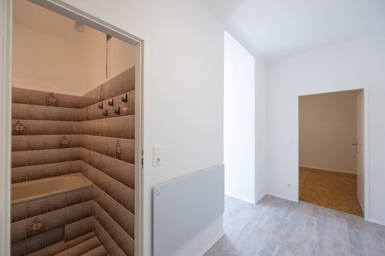 Blick vom Flur in ein kleines Badezimmer mit Badewanne und braunen Fliesen an den Wänden.