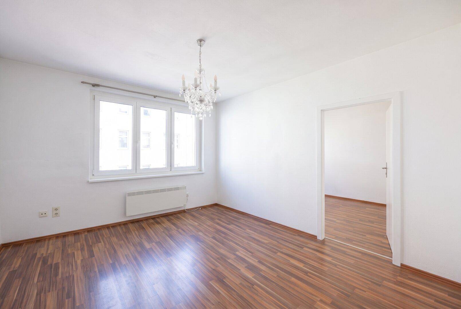Helles, leeres Zimmer mit Parkettboden, weißen Wänden und einem eleganten Kronleuchter.