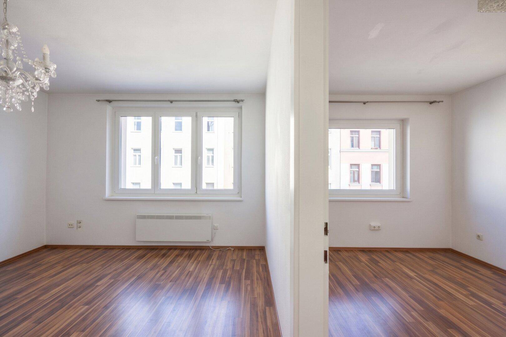Zwei helle, leere Räume mit Parkettboden und Fenstern, getrennt durch eine Wand.