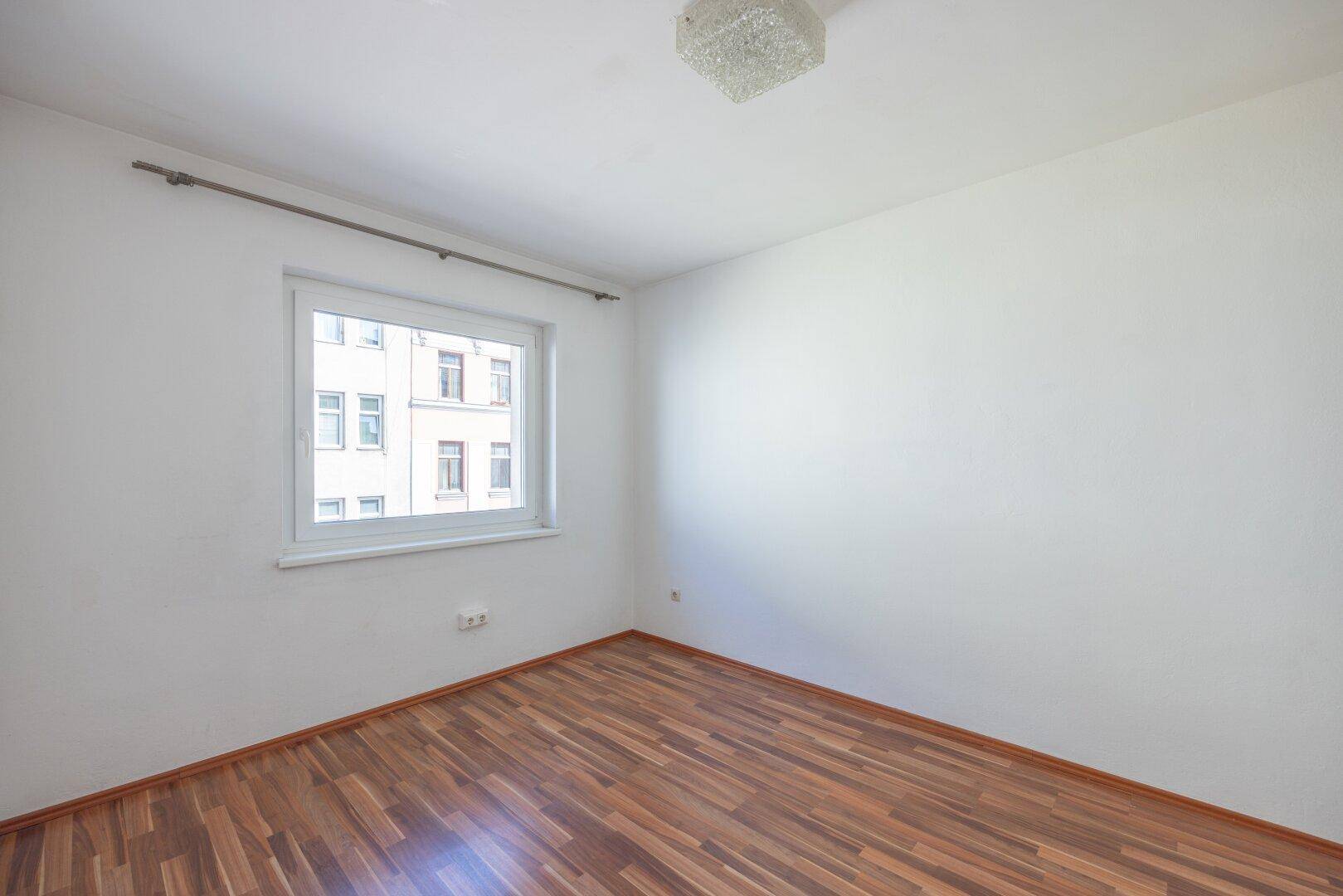 Schlichtes, helles Zimmer mit Parkettboden, weißen Wänden und einem Fenster.