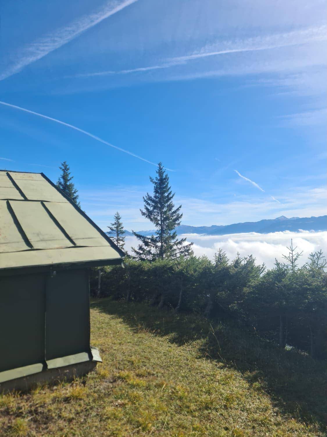 Kleine grüne Hütte auf einem Hügel mit Blick über eine Wolkendecke und blauen Himmel.