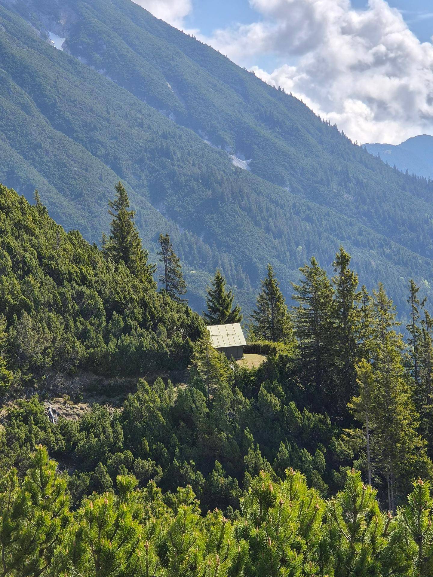 Eine kleine Hütte ist eingebettet in eine üppige, grüne Berglandschaft unter einem bewölkten Himmel.