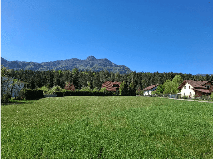 Weitläufige grüne Wiese mit Bergblick und umliegenden Häusern an einem sonnigen Tag.