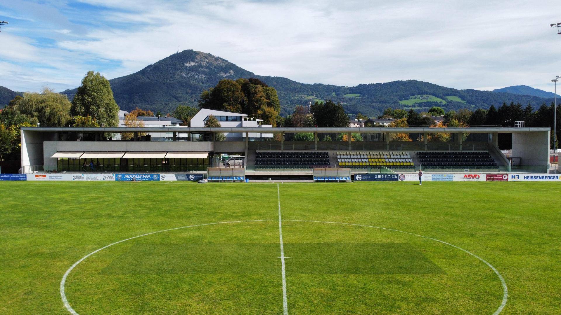 Weitläufige Ansicht eines Sportstadions mit grüner Rasenfläche und Tribünen vor einer Bergkulisse.