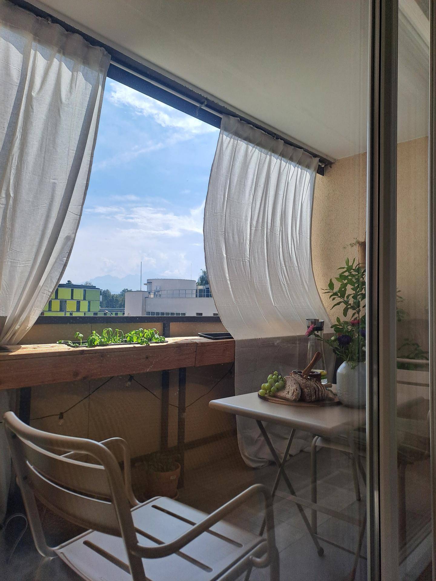 Heller Balkon mit kleinem Tisch, Stühlen und Vorhängen, ideal für Mahlzeiten im Freien.