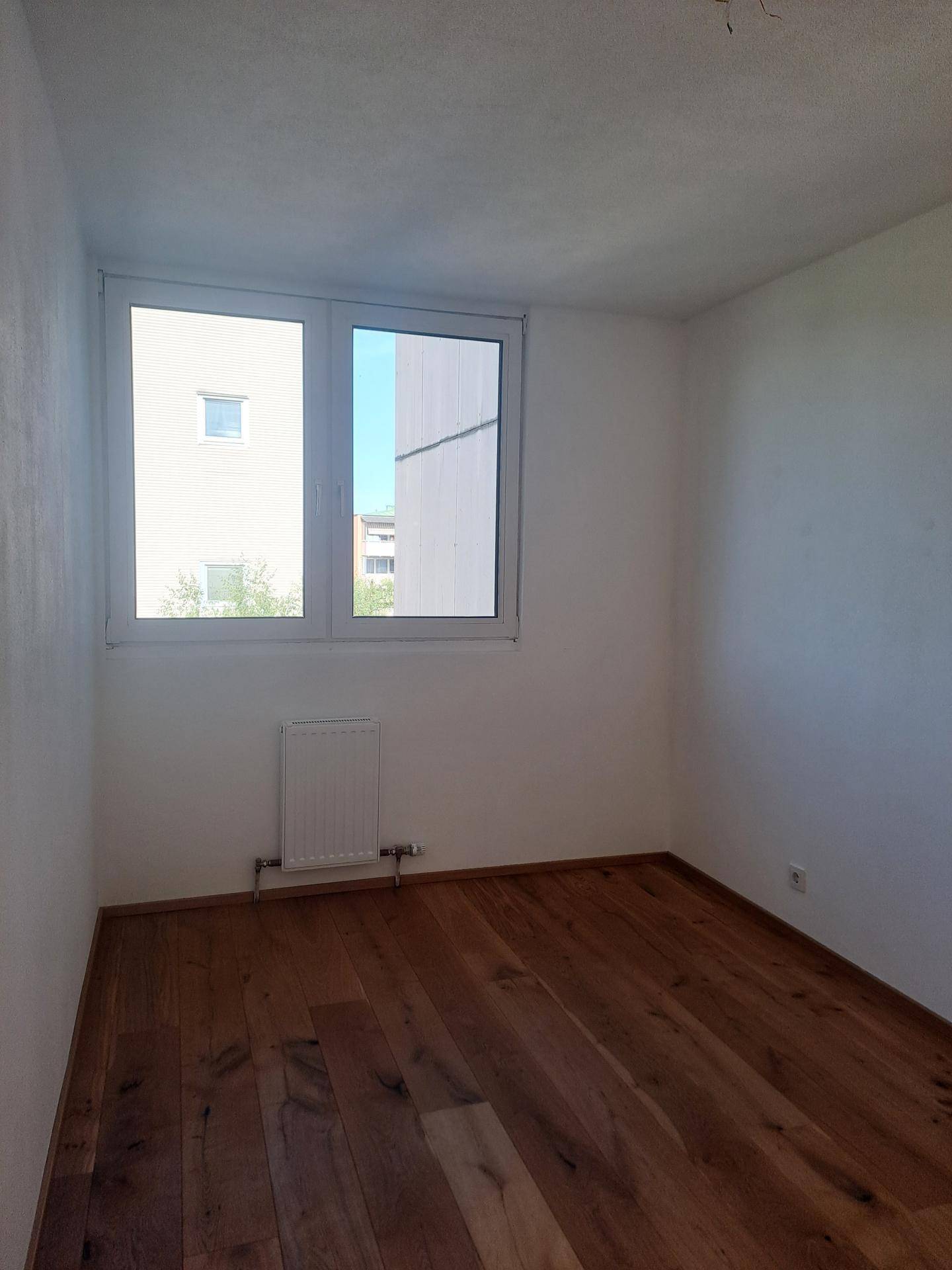 Leeres Zimmer mit Holzboden, weiß gestrichenen Wänden und großem Fenster.