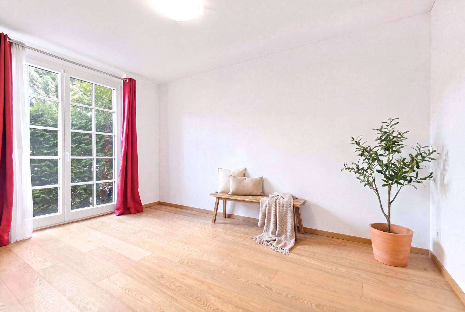Helles Zimmer mit Holzboden, großen Fenstern und roten Vorhängen, mit Blick ins Grüne.