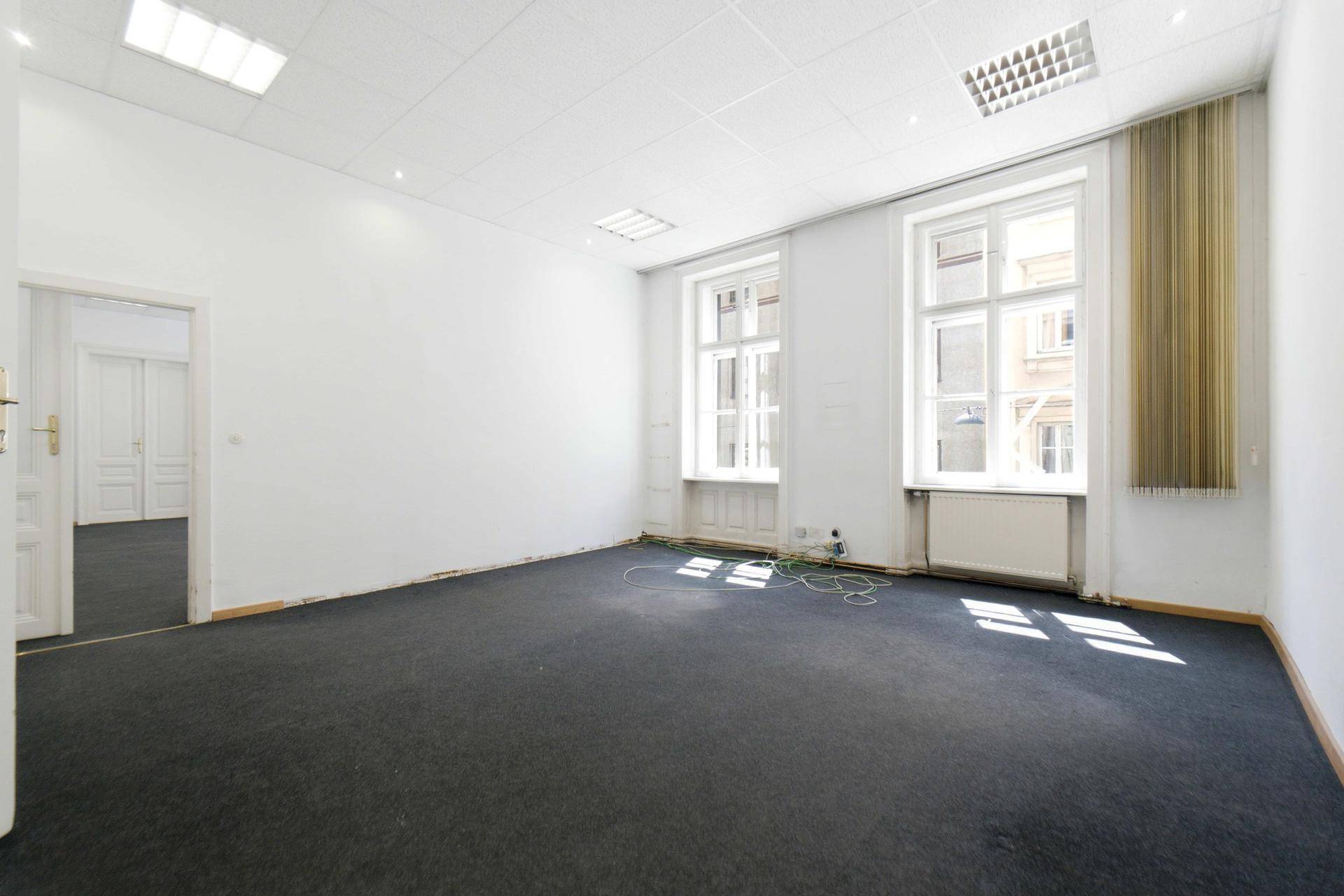 Heller Raum mit dunklem Teppichboden und zwei Fenstern, mit Zugang zu einem weiteren Zimmer.