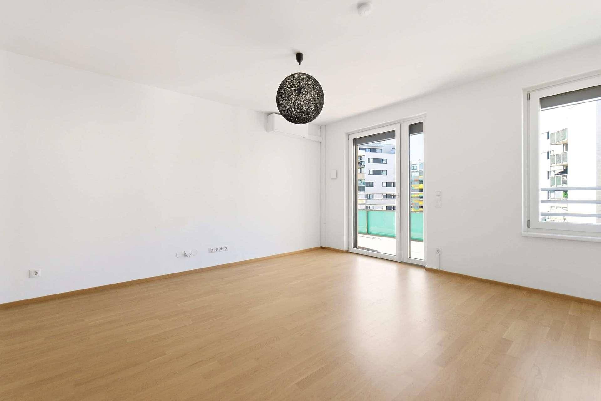 Leeres Wohnzimmer mit hellem Holzboden, großen Fenstern und Zugang zum Balkon.