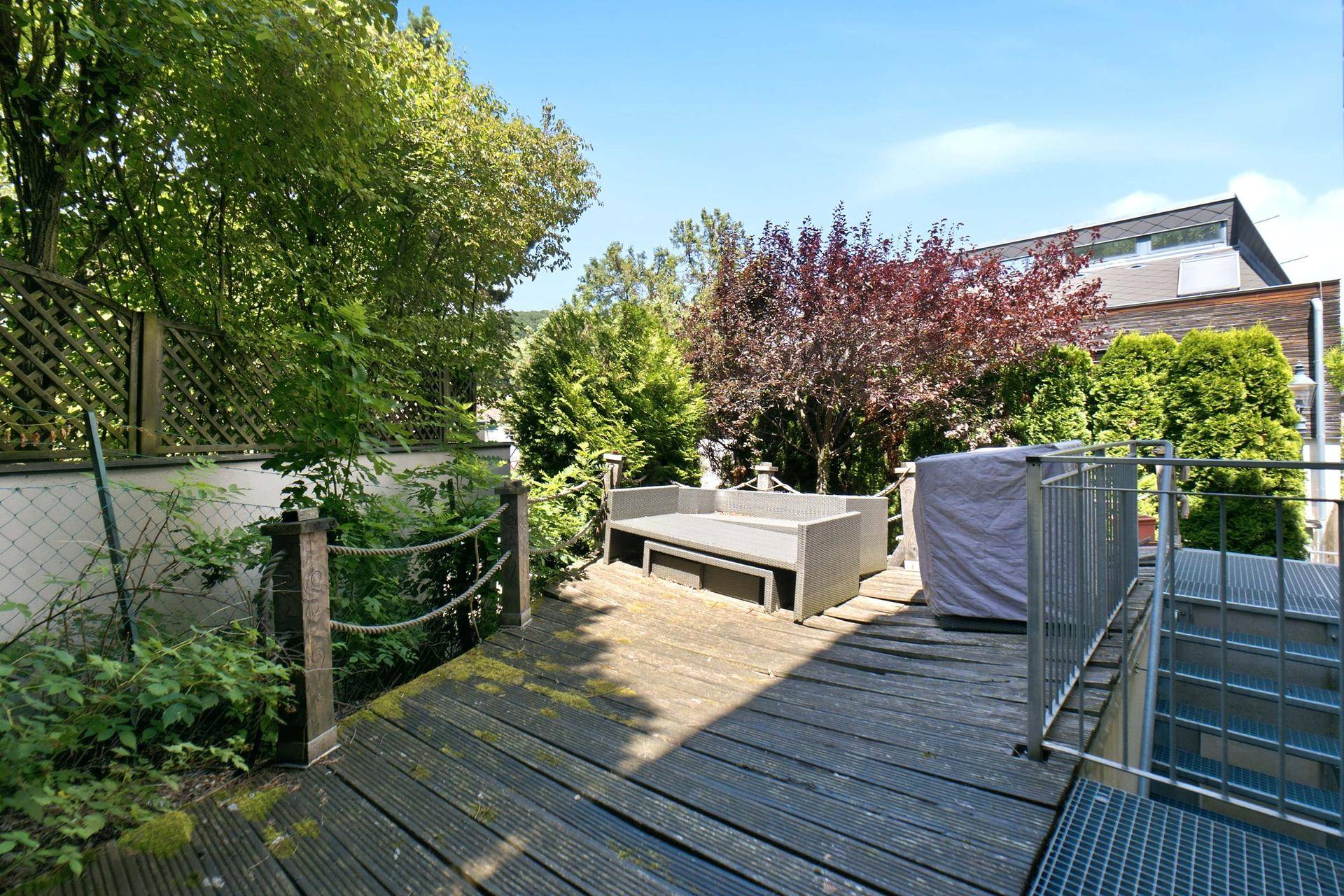 Holzterrasse mit bequemer Outdoor-Sitzgelegenheit, umgeben von üppiger Vegetation.