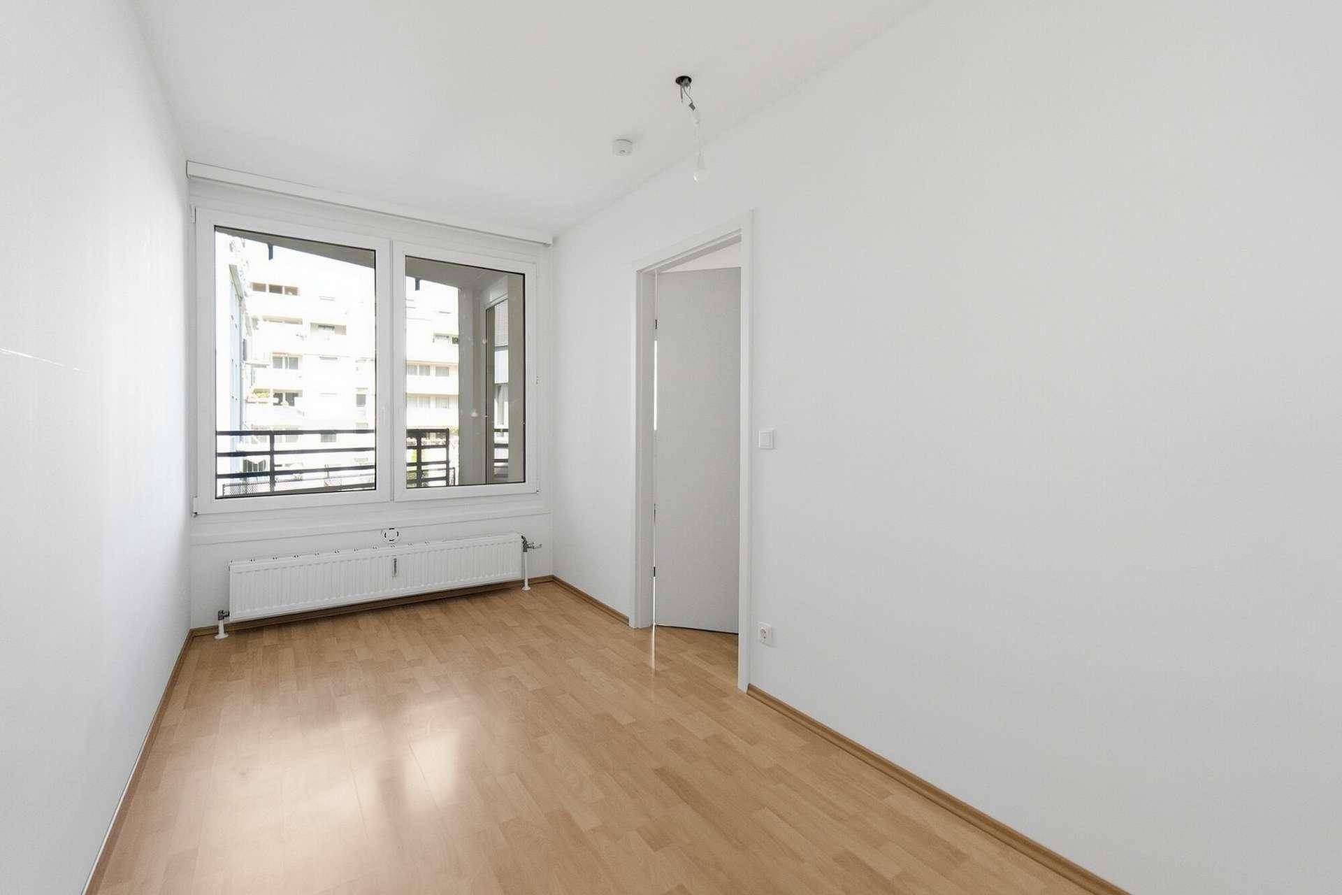Helles, leeres Zimmer mit Parkettboden, weißen Wänden und einem Fenster mit Blick ins Freie.