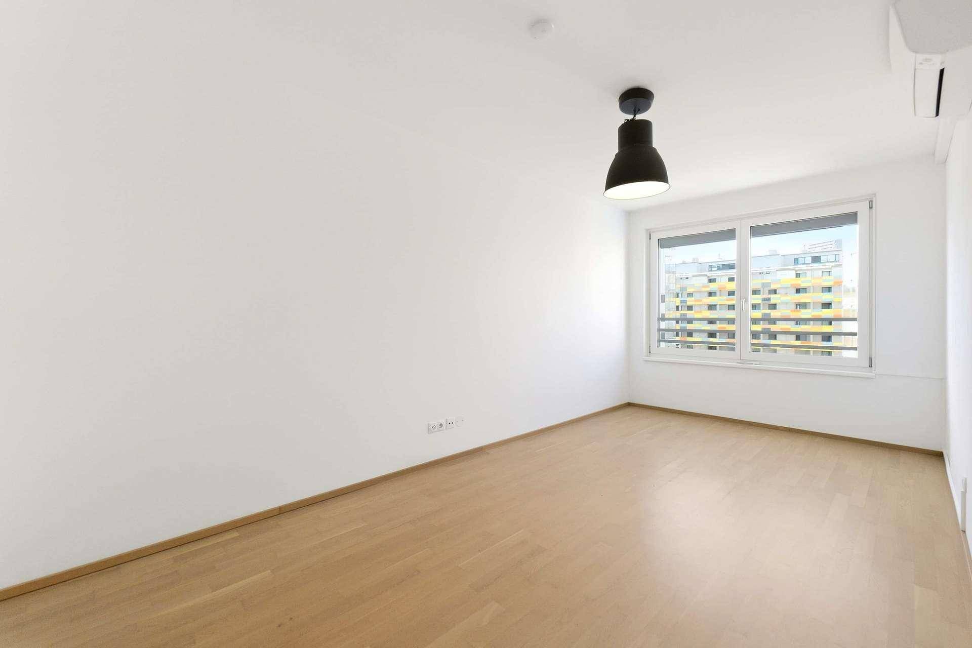 Leeres Schlafzimmer mit hellem Holzboden, großen Fenstern und Klimaanlage.