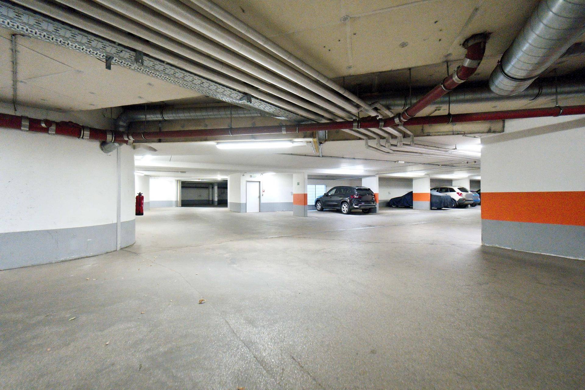 Weitläufige Tiefgarage mit Betonboden, weißen Wänden und orangefarbenen Markierungen an den Säulen.