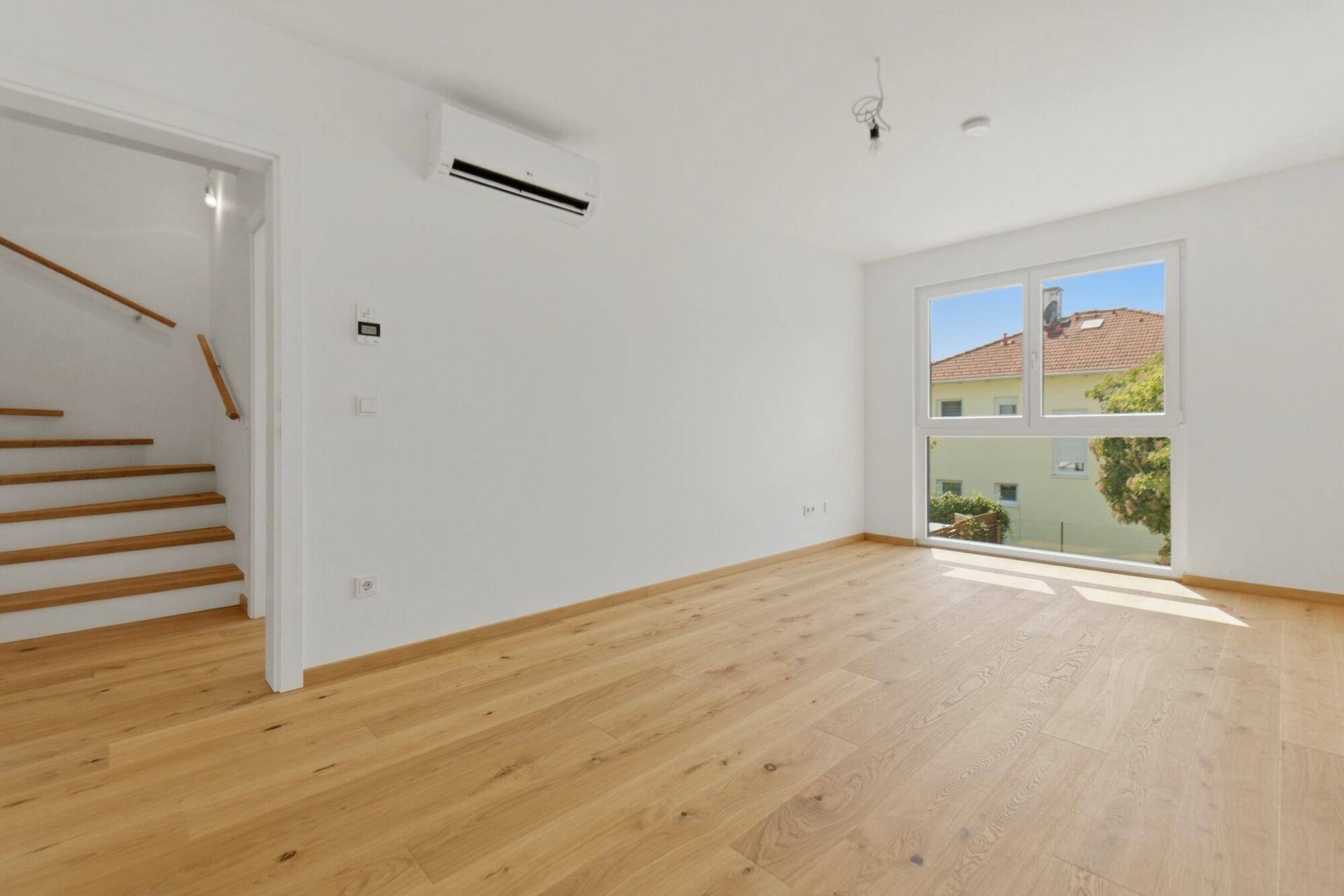 Heller Wohnraum mit Holzboden, großen Fenstern und Klimaanlage, angrenzend an eine Holztreppe.
