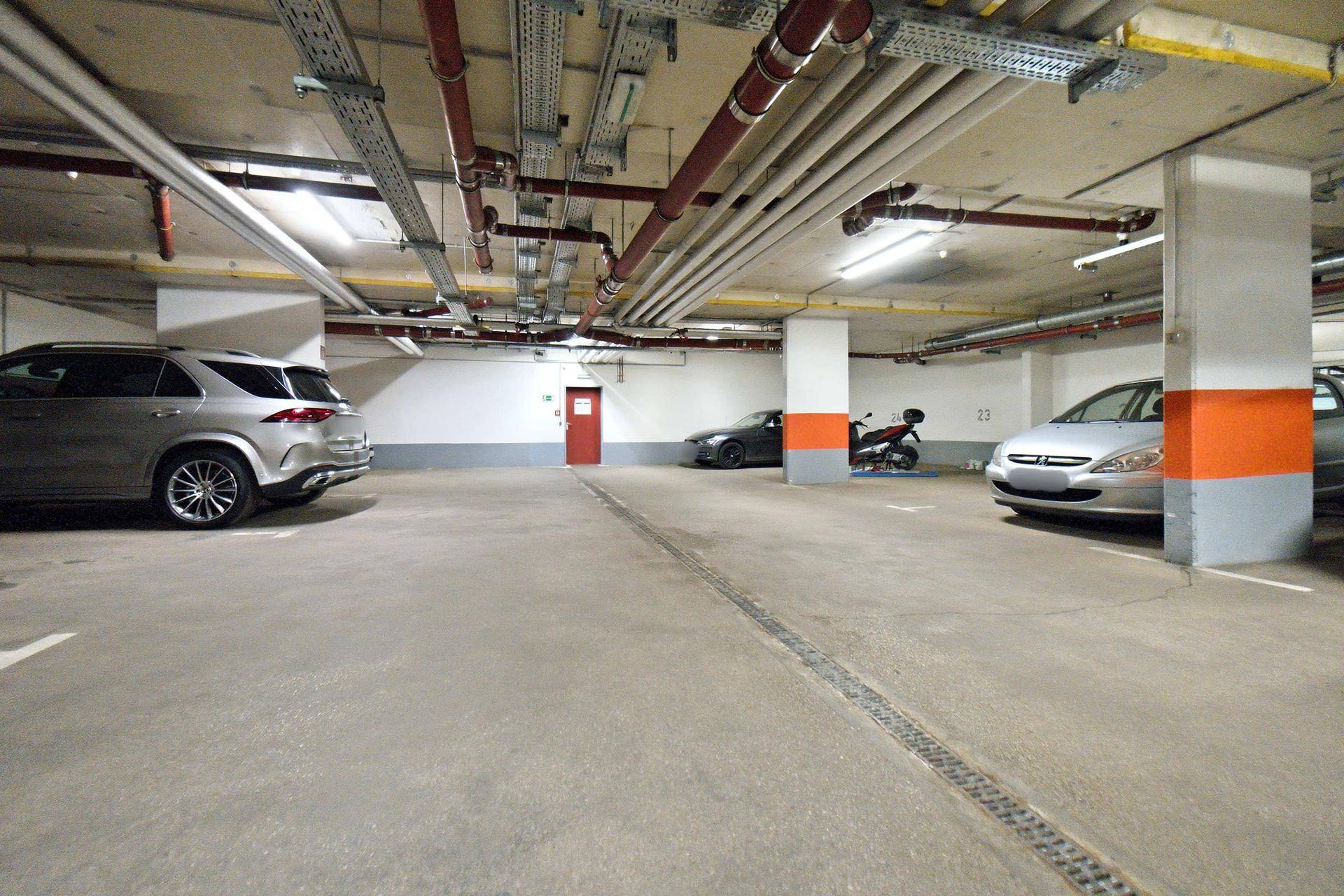 Tiefgarage mit Betonboden, orangefarbenen Säulen und sichtbaren Rohren an der Decke.