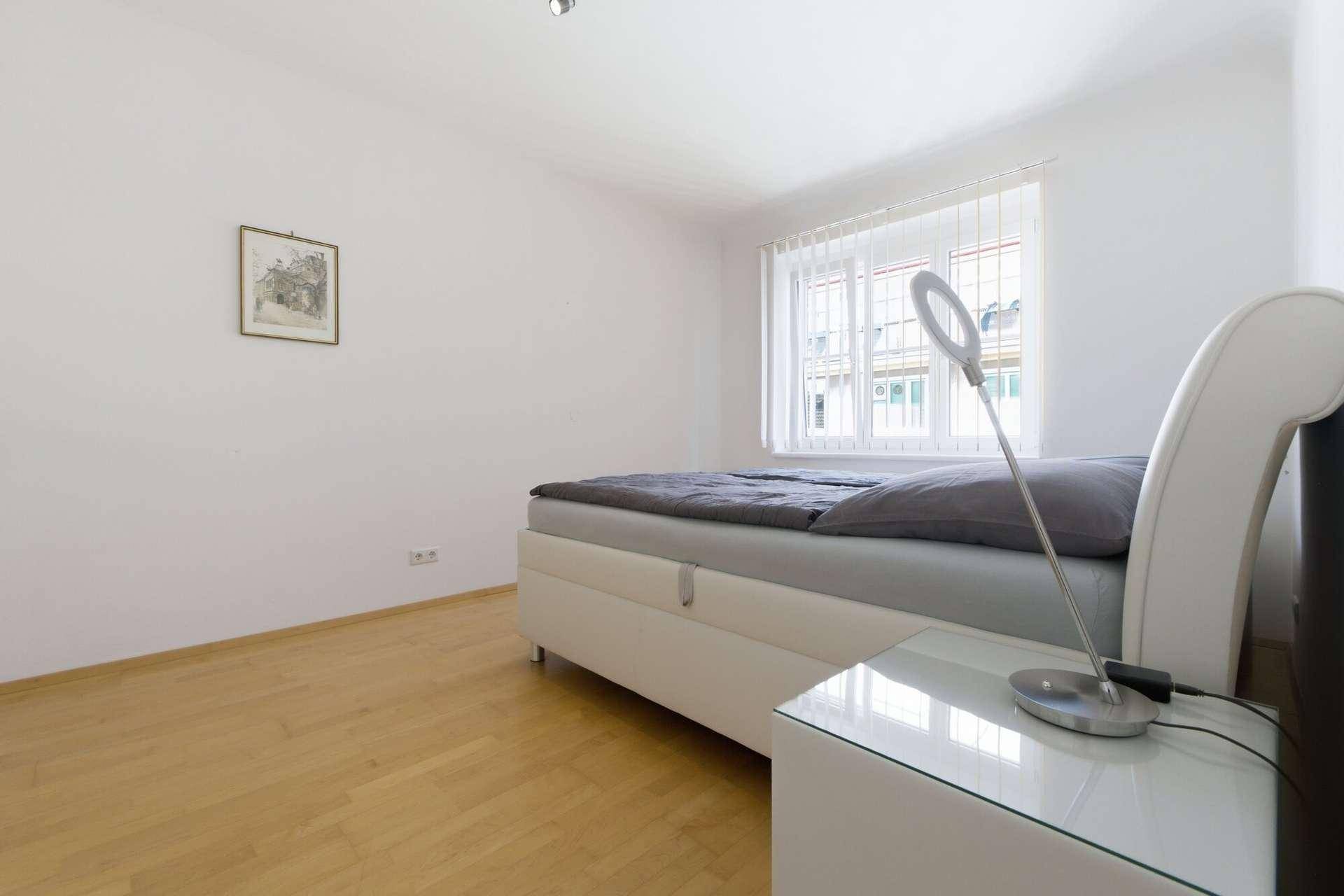 Schlafzimmer mit weißem Bett, Nachttisch und Fenster, das viel Licht hereinlässt.