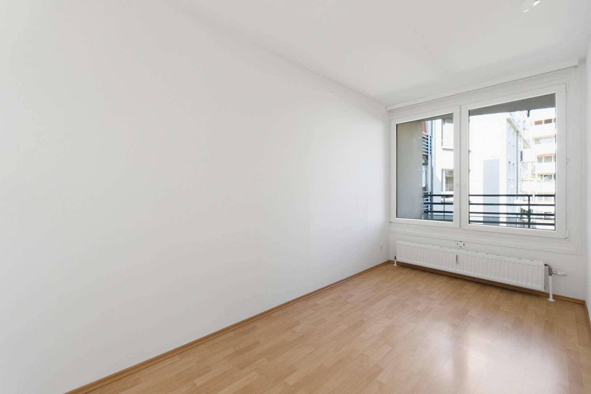 Leeres Zimmer mit Parkettboden, weißen Wänden und großem Fenster mit Blick nach draußen.
