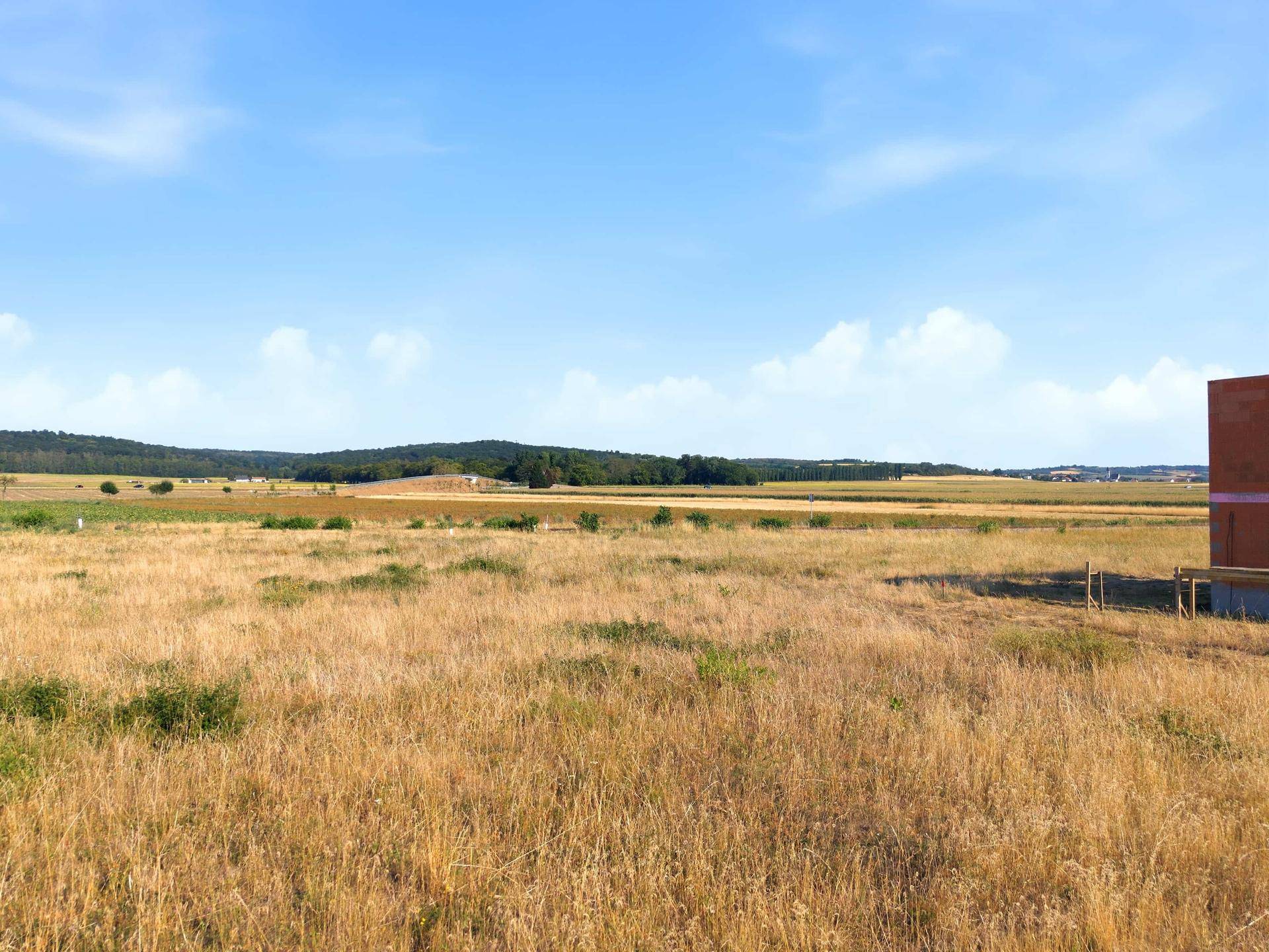 Weites, trockenes Feld unter blauem Himmel mit einem verschwommenen Gebäude am rechten Bildrand.