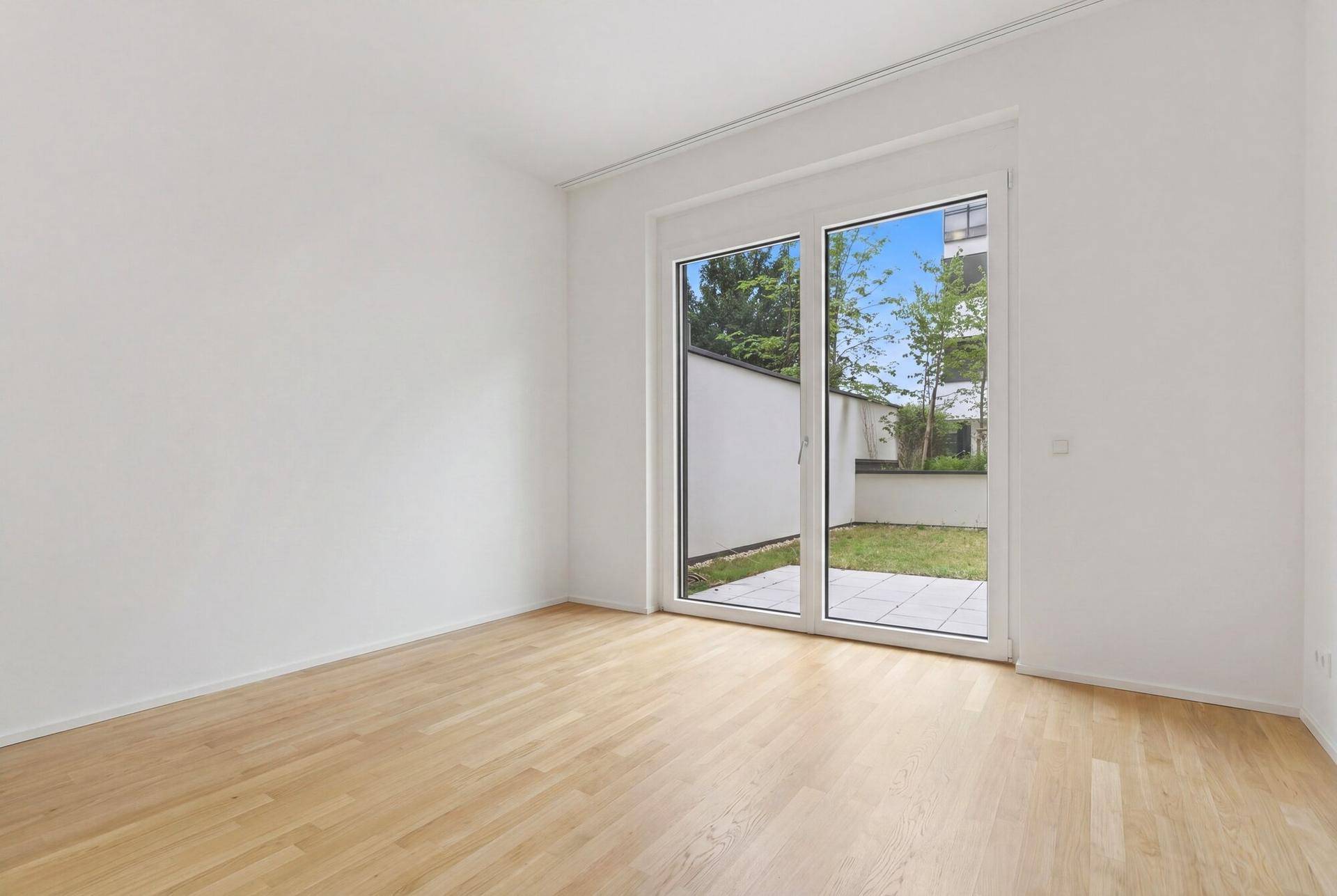 Helles, leeres Zimmer mit Parkettboden und Zugang zu einer kleinen Terrasse und Garten.