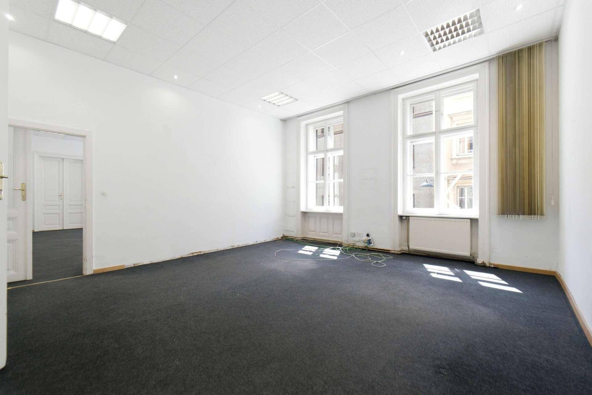 Heller, leerer Raum mit Teppichboden, großen Fenstern und Zugang zu einem weiteren Zimmer.