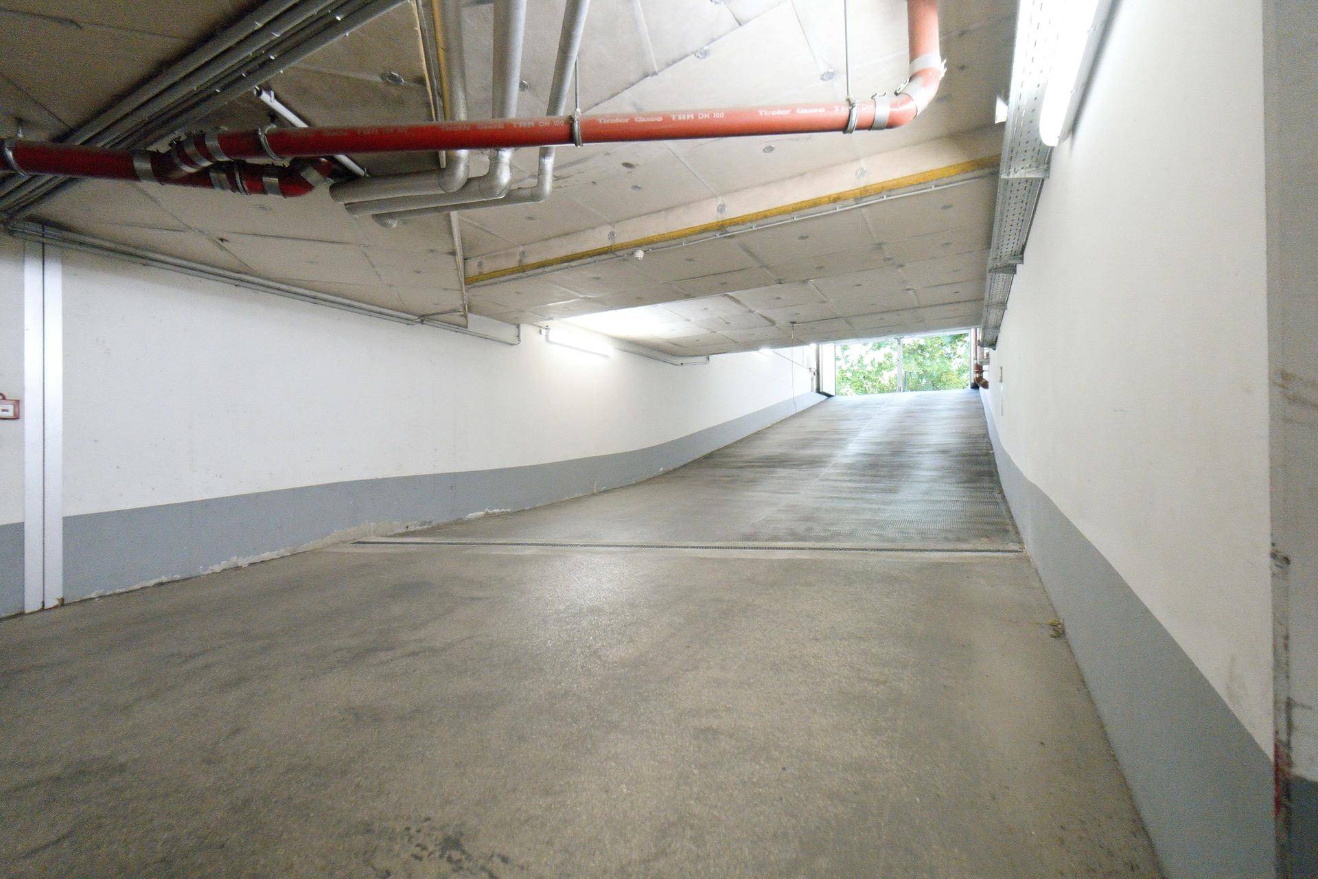 Zufahrtsrampe zur Tiefgarage mit Betonboden und sichtbaren Versorgungsleitungen an der Decke.