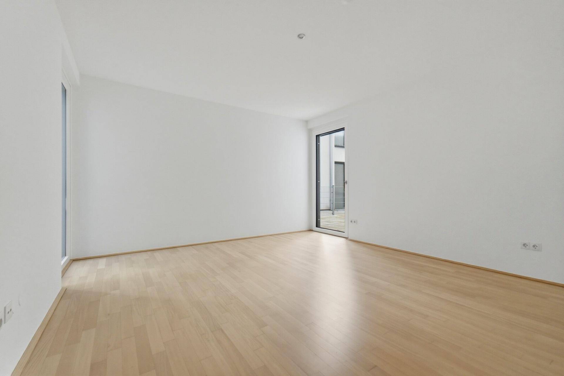 Helles Zimmer mit Holzboden und Zugang zu einem Außenbereich durch eine Fenstertür.