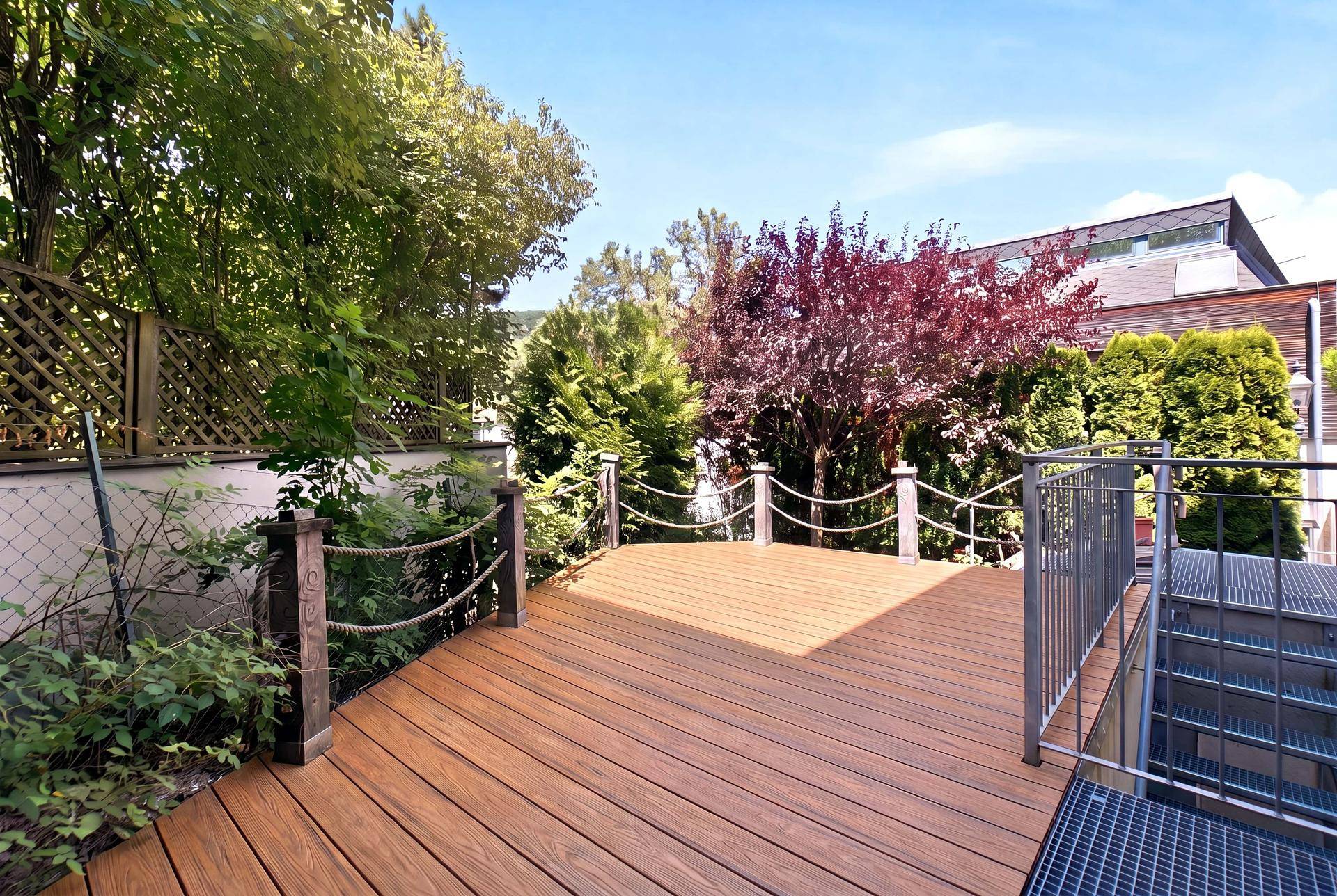 Holzterrasse mit Seilgeländer und Treppenzugang, umgeben von Bäumen und Sträuchern.