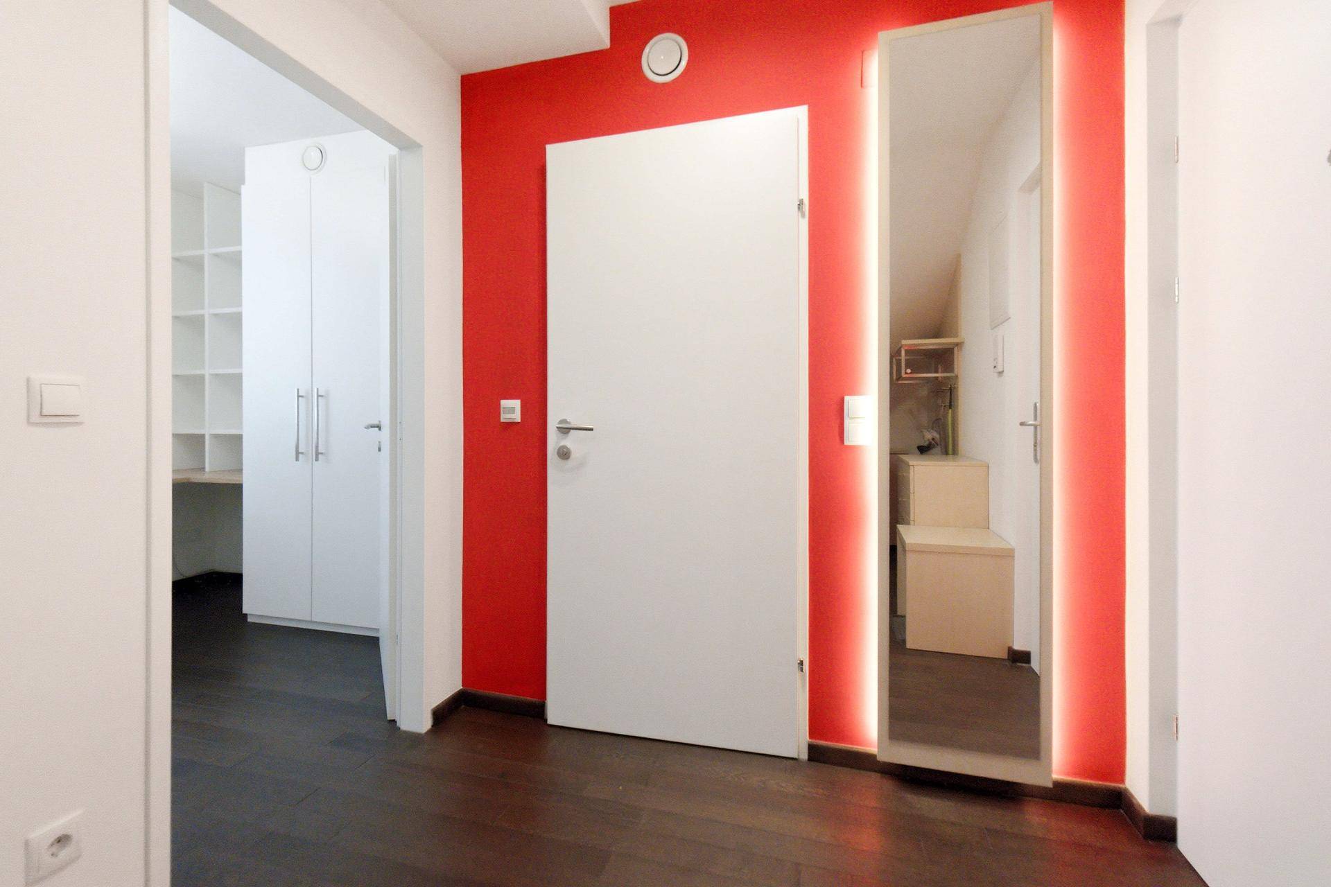 Heller Flur mit roter Akzentwand und beleuchtetem Spiegel, führt zu den Schlafzimmern.