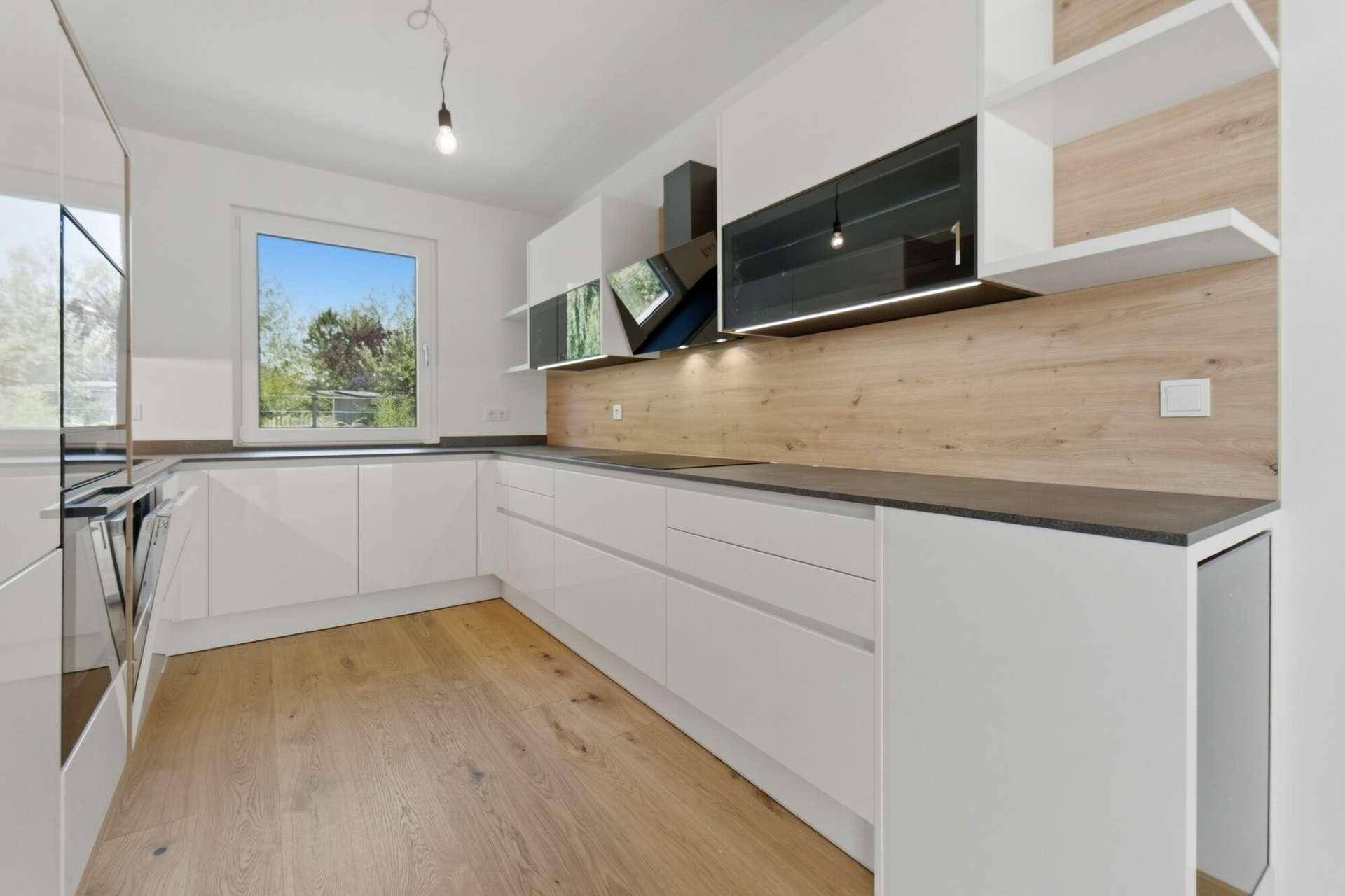 Moderne weiße Küche mit Holzdetails, Einbaugeräten und Fenster mit Blick ins Grüne.