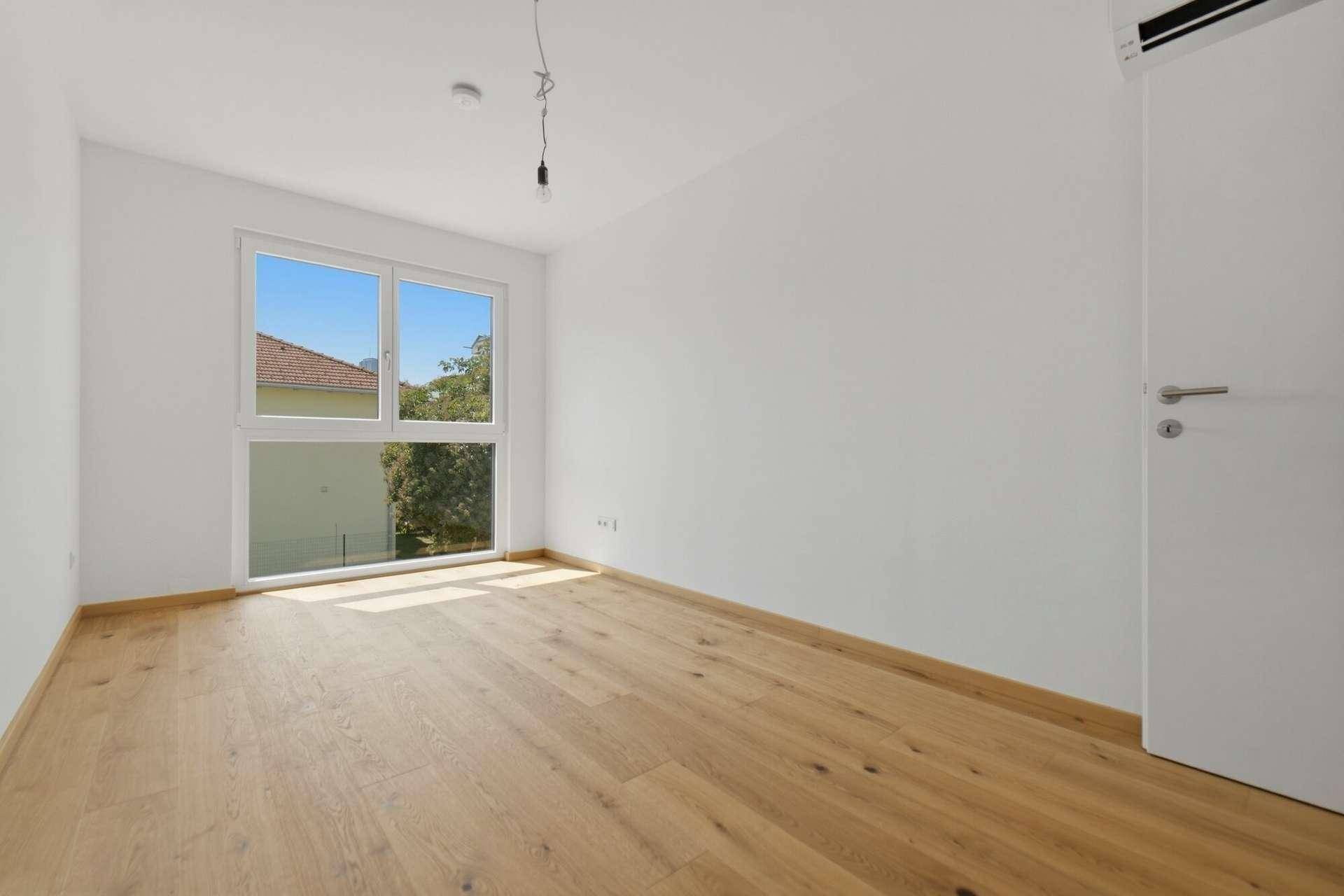 Lichtdurchflutetes Schlafzimmer mit Holzboden und großem Fenster, das einen Blick ins Grüne bietet.