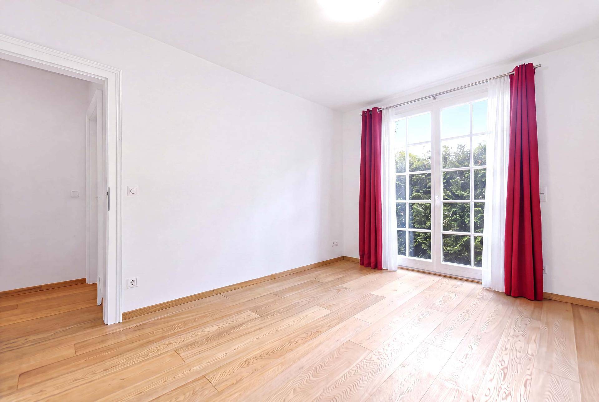 Geräumiges Zimmer mit Holzboden, großen Fenstern und roten Vorhängen, viel Tageslicht.