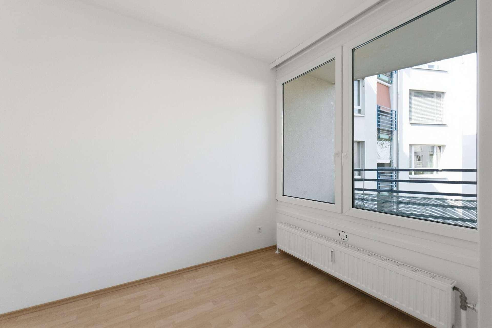 Leeres Zimmer mit Parkettboden, weißen Wänden und großem Fenster mit Blick auf die Umgebung.