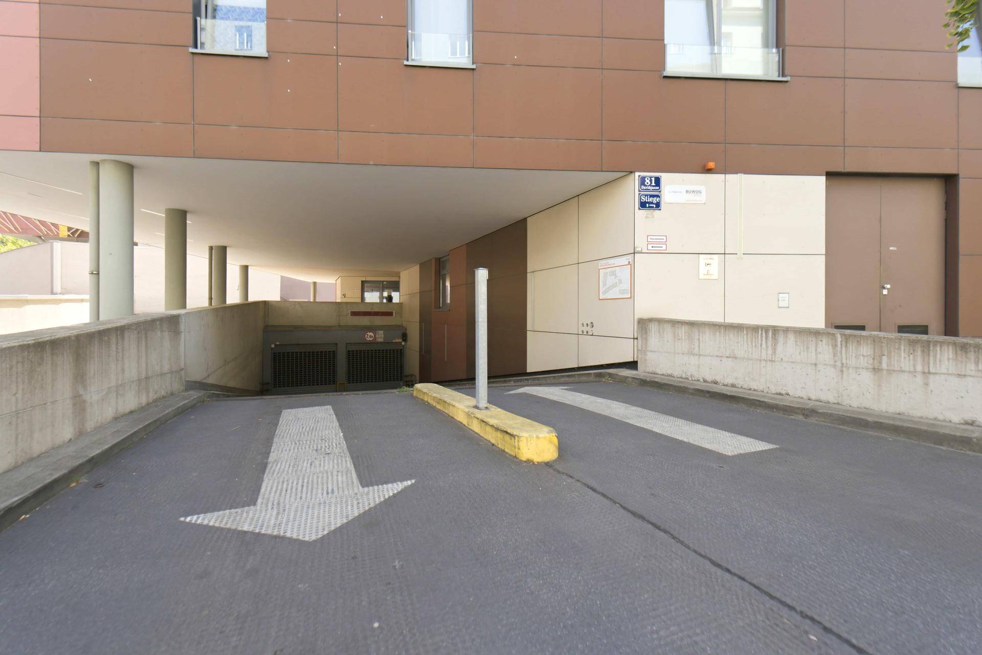 Einfahrt zu einer Tiefgarage unter einem modernen Wohngebäude mit brauner Fassade und Pfeilmarkierungen.