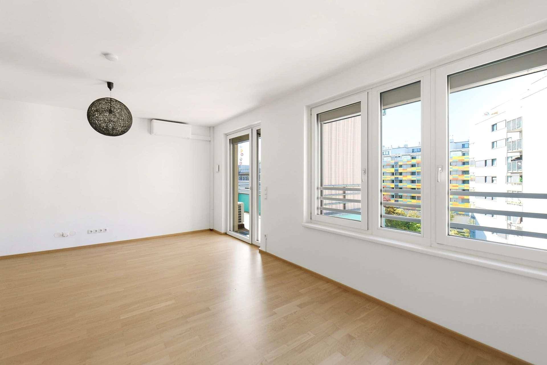 Heller Raum mit Holzboden, großen Fenstern und Zugang zu einem Balkon.