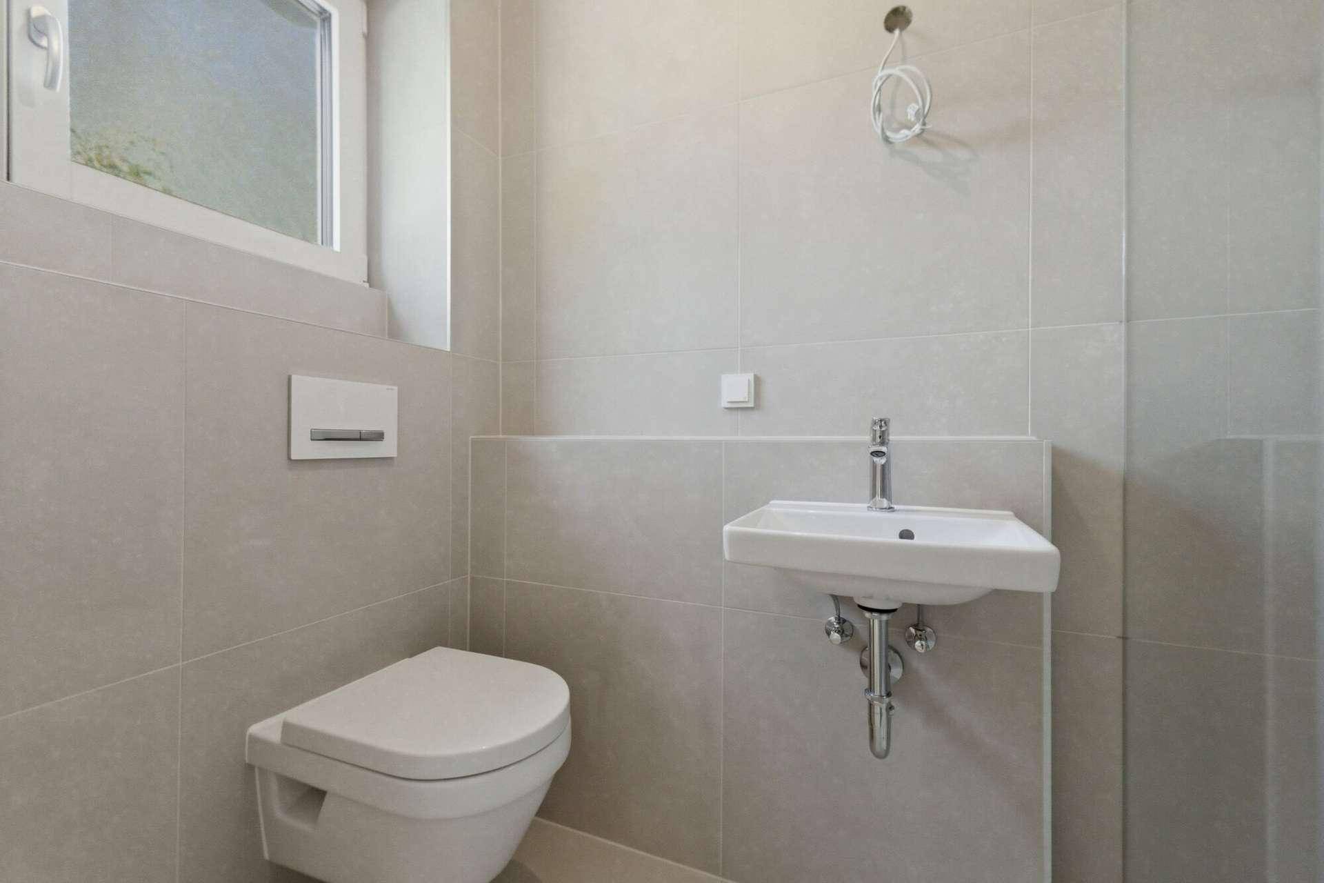 Modernes Gäste-WC mit Hänge-WC, Waschbecken und Fenster, hell gefliest.
