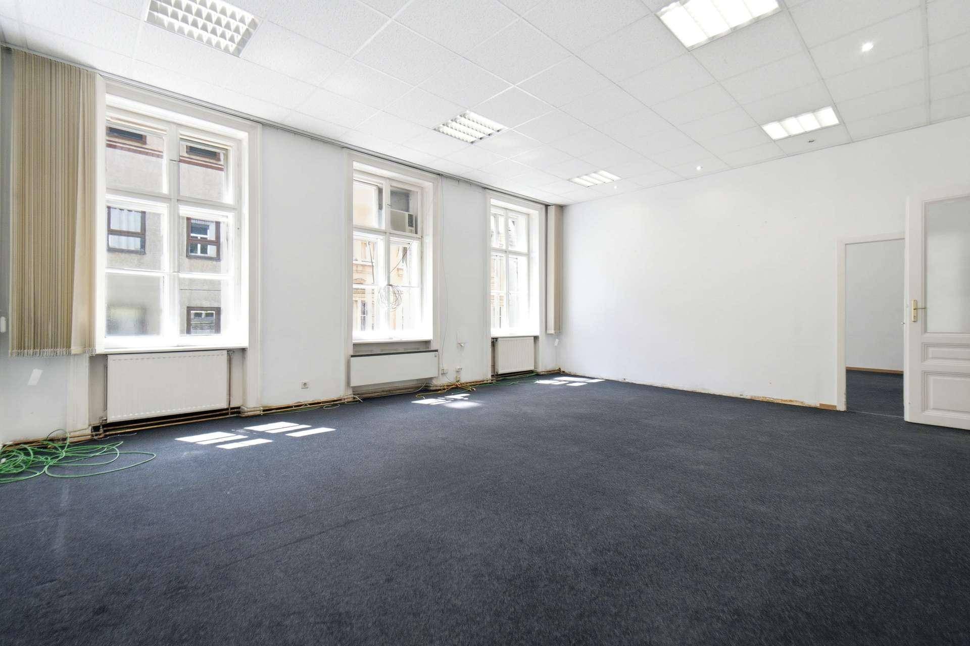 Großer, leerer Raum mit dunklem Teppichboden, weißen Wänden und drei Fenstern, viel Tageslicht.
