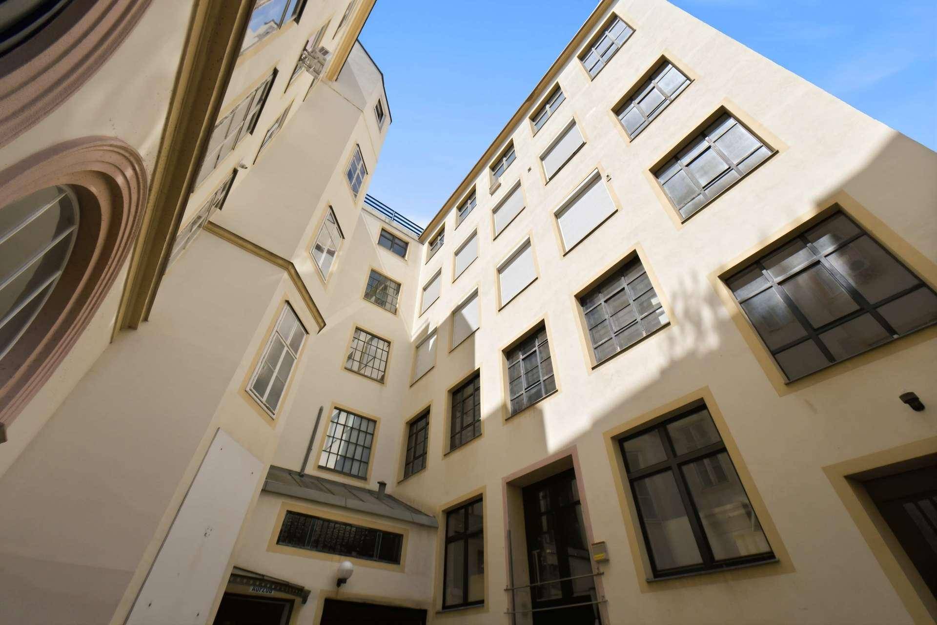 Blick in den Innenhof eines Gebäudes mit mehreren Fenstern und heller Fassade unter blauem Himmel.