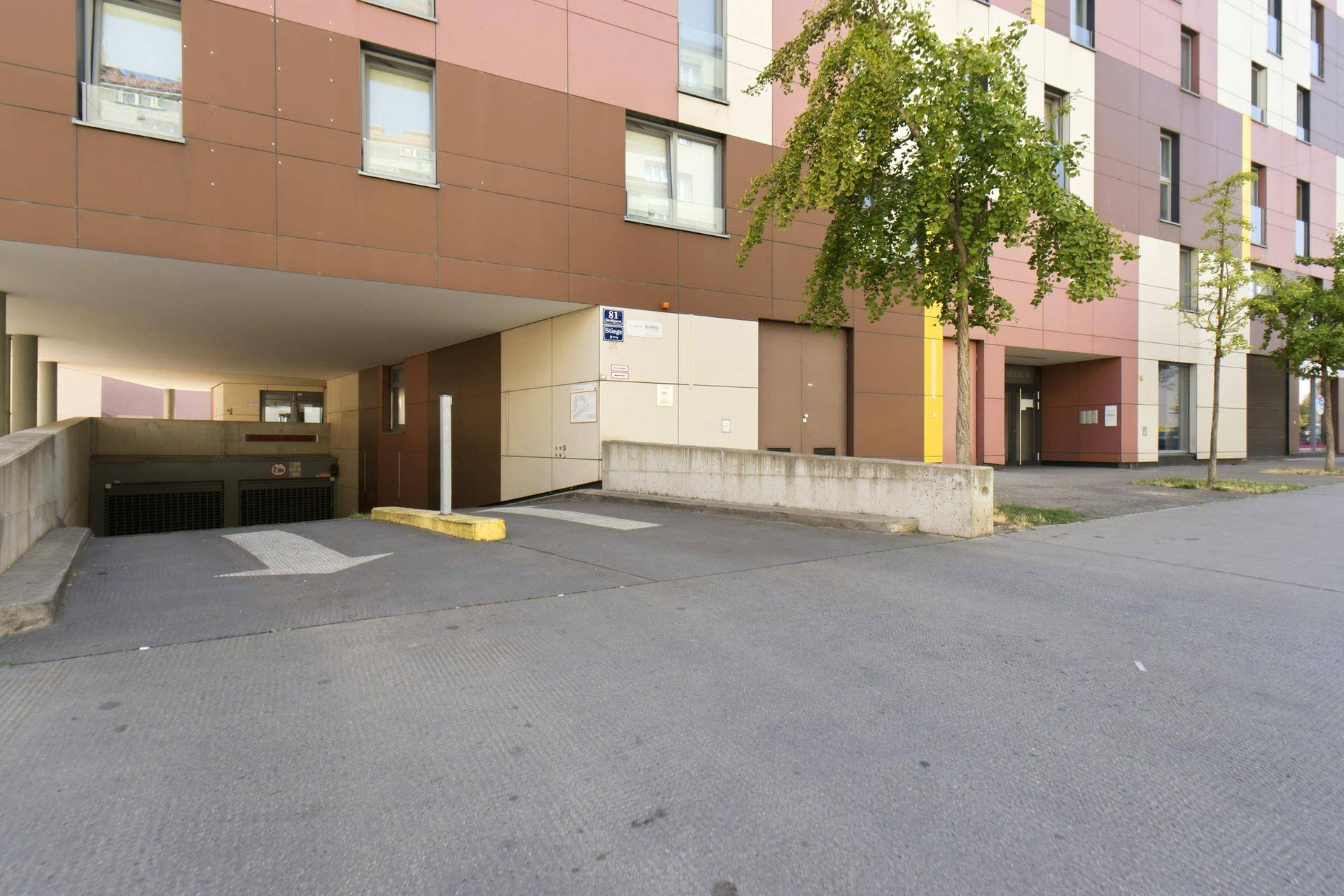Außenansicht der Tiefgarageneinfahrt eines modernen Wohngebäudes mit Baum und brauner Fassade.