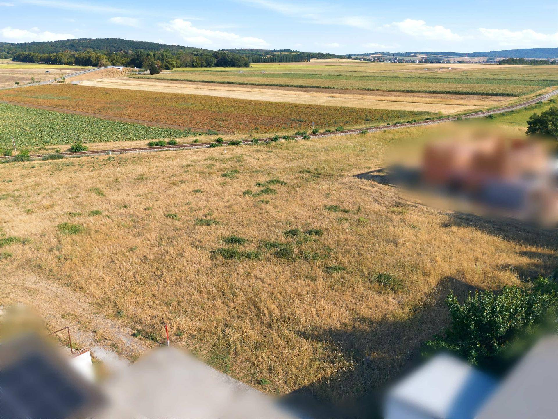 Luftaufnahme eines weiten Feldes mit ländlicher Umgebung und verschwommenen Gebäuden im Hintergrund unter blauem Himmel.