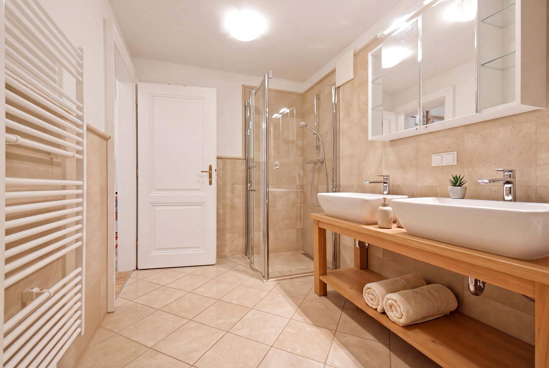 Modernes Badezimmer mit Doppelwaschbecken, Dusche und beheiztem Handtuchhalter, hell gefliest.