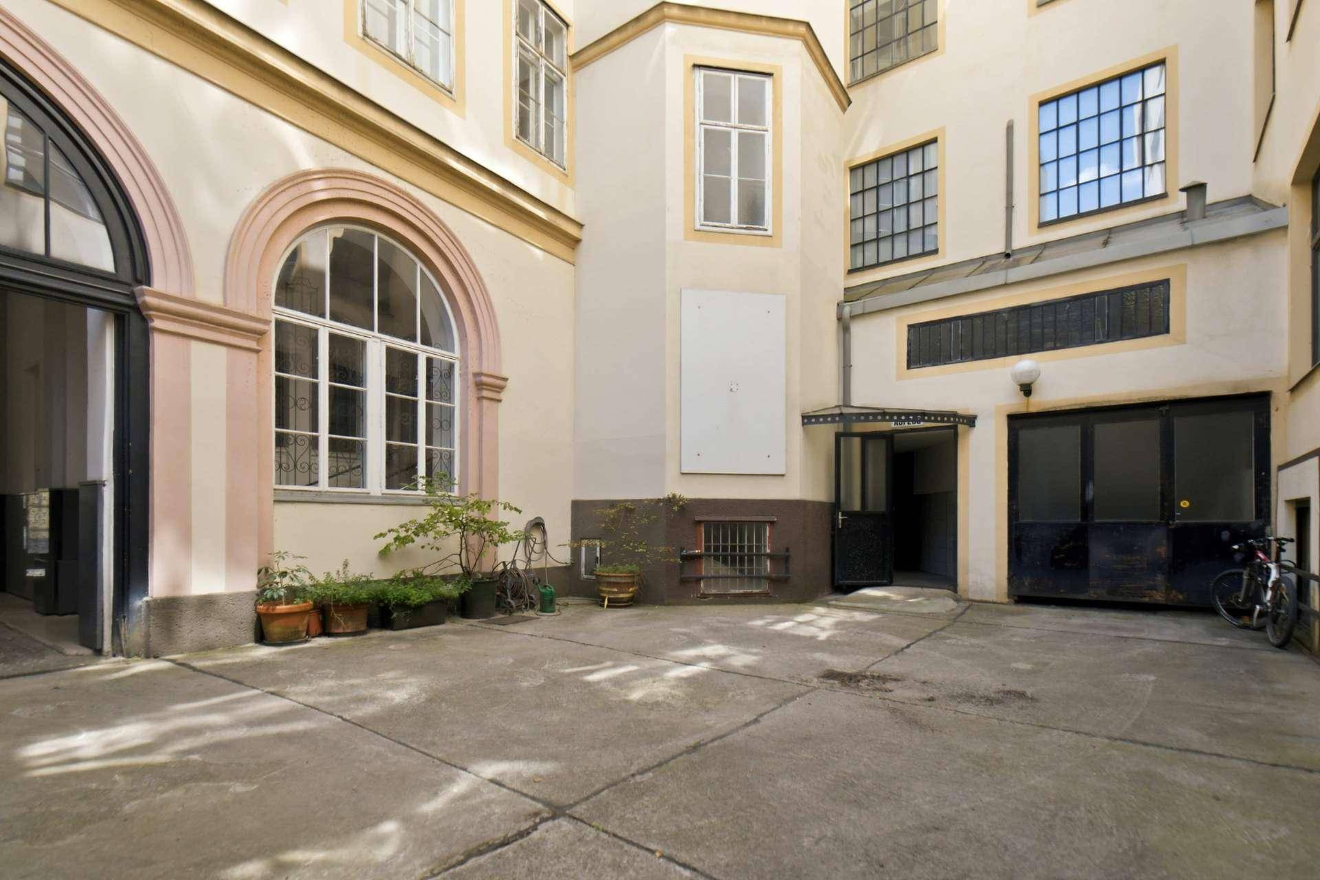 Heller Innenhof eines Gebäudes mit Betonboden, verschiedenen Fenstern und Pflanzen, ruhige Atmosphäre.