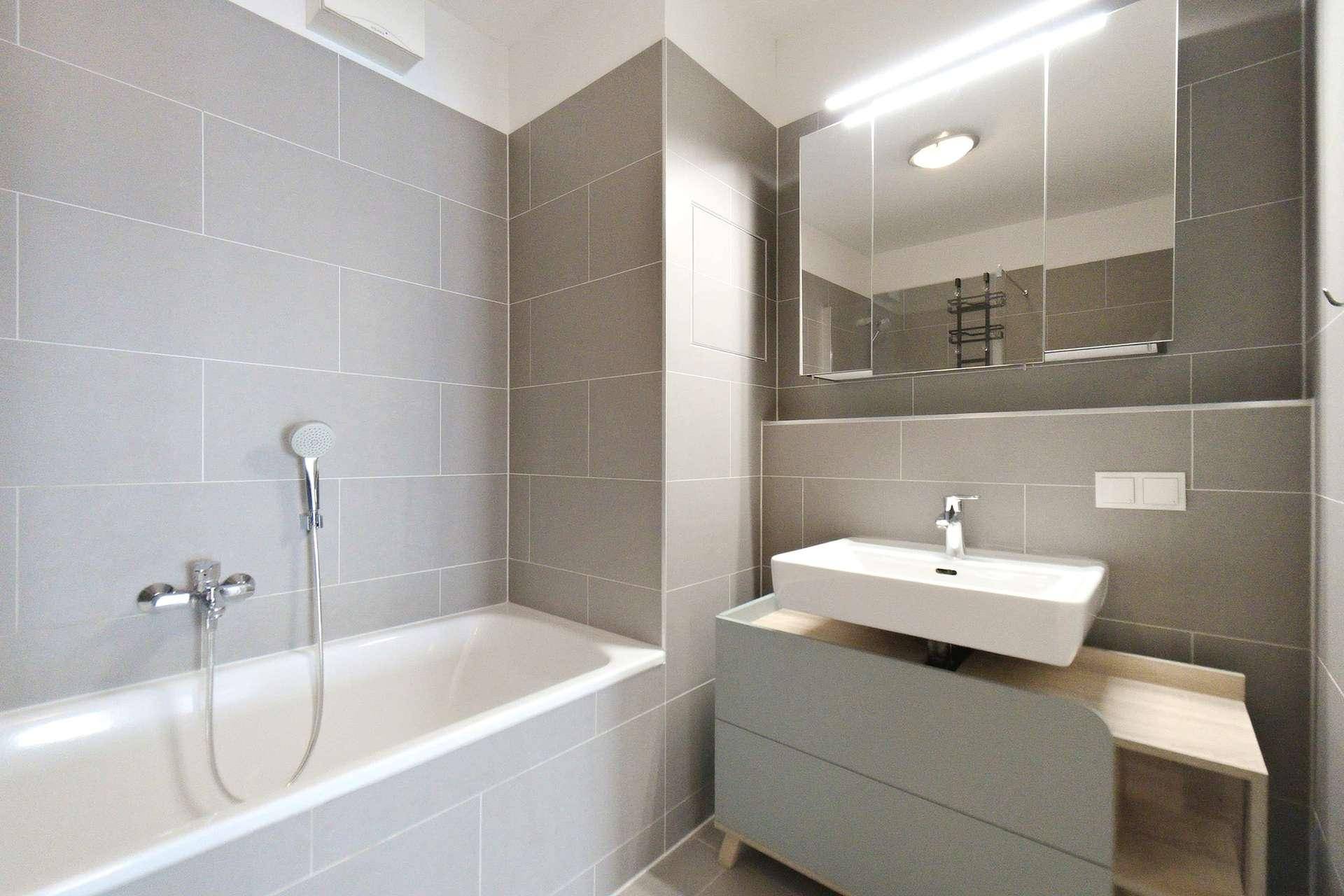 Zeitgemäßes Badezimmer mit Badewanne, Waschtisch, Spiegelschrank und grauen Wandfliesen.