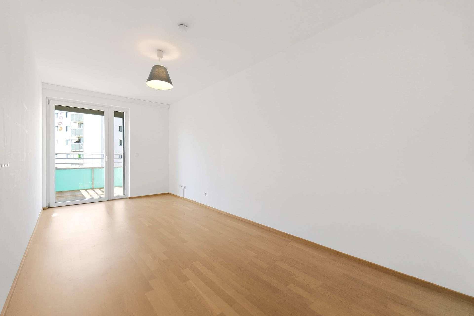Heller Raum mit Holzboden, großen Fenstern und Zugang zum Balkon.