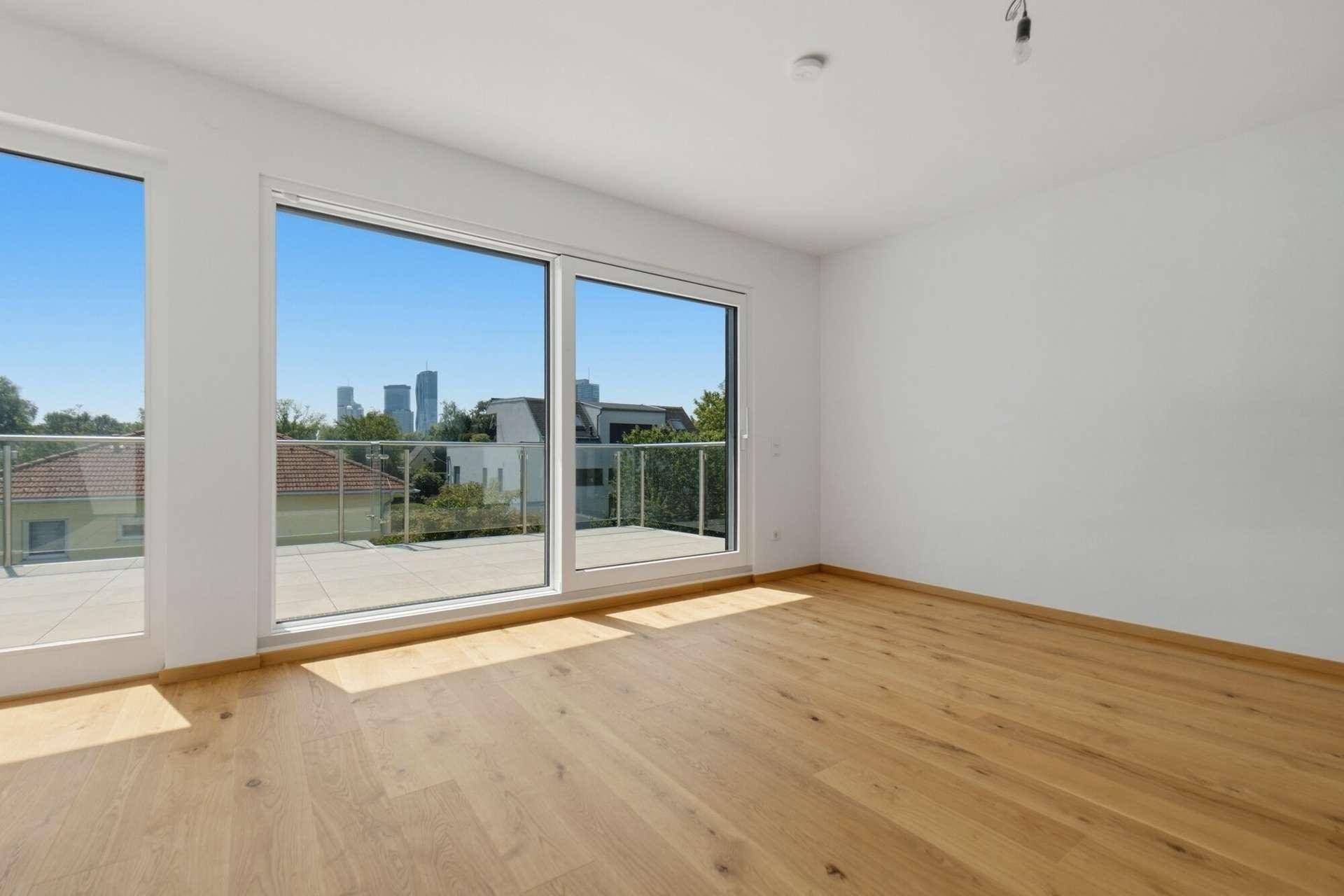 Heller Raum mit Holzboden, großen Fenstern, Terrassenzugang und Stadtblick in der Ferne.