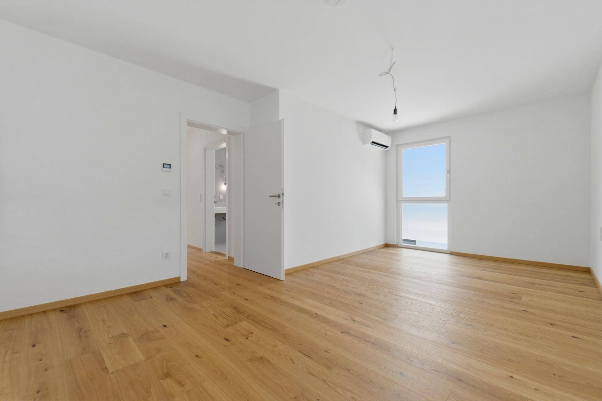 Geräumiges Zimmer mit Holzboden, Fenster und Klimaanlage, mit Zugang zu einem weiteren Raum.