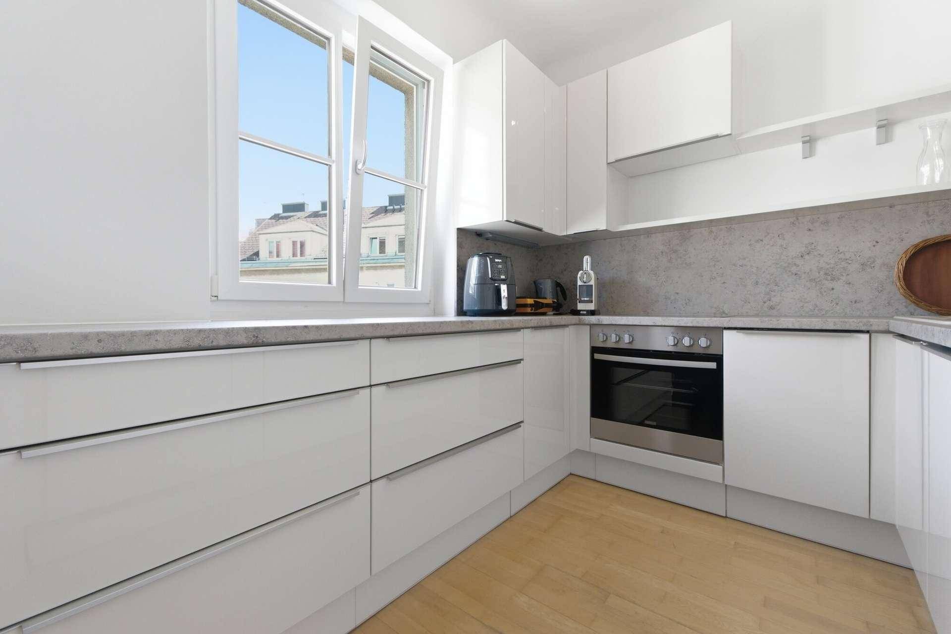 Moderne weiße Küche mit glänzenden Fronten, integriertem Backofen und Fensterblick.