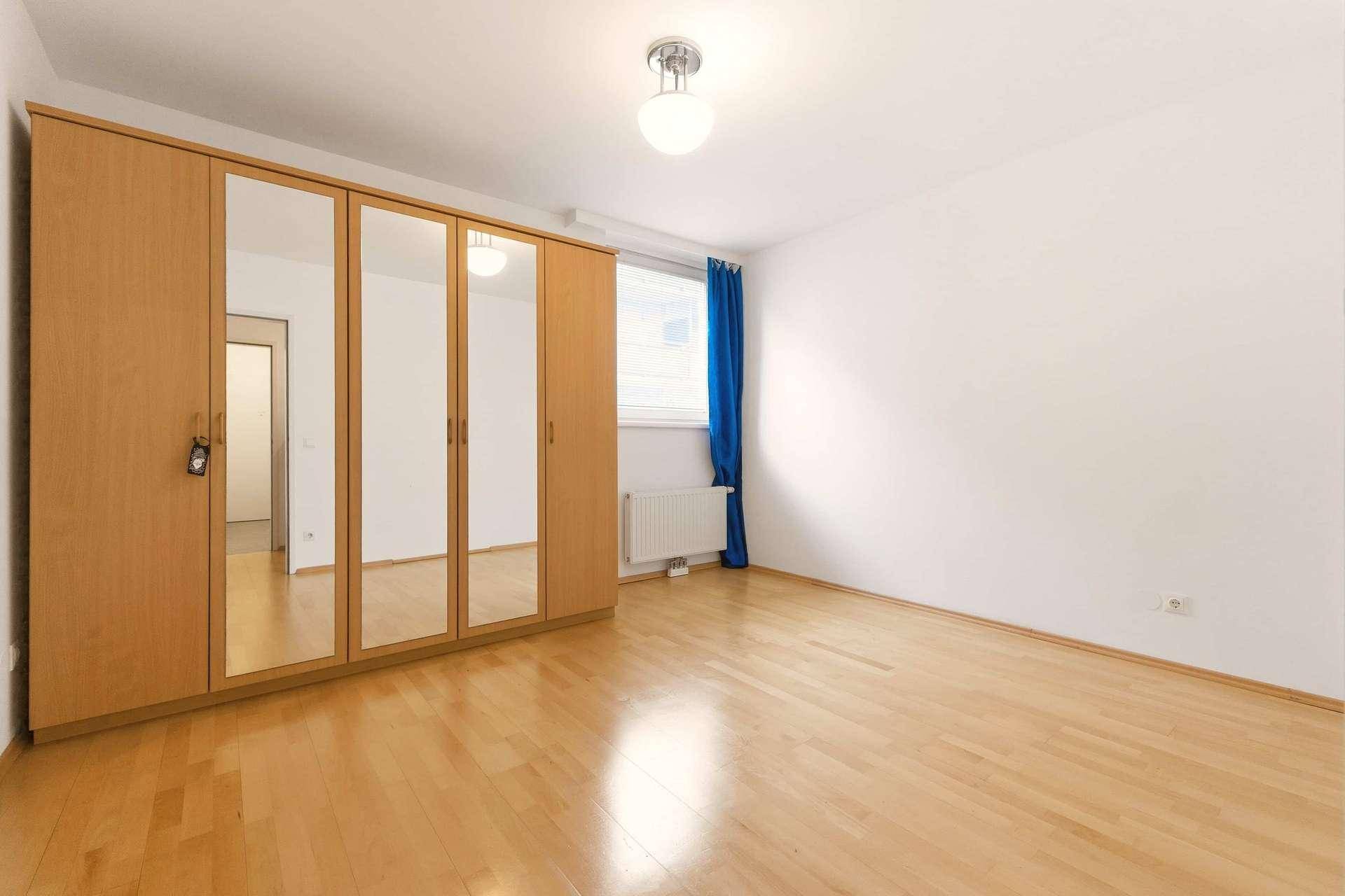 Schlafzimmer mit hellem Parkettboden, großem Spiegelschrank und Fenster mit blauen Vorhängen.