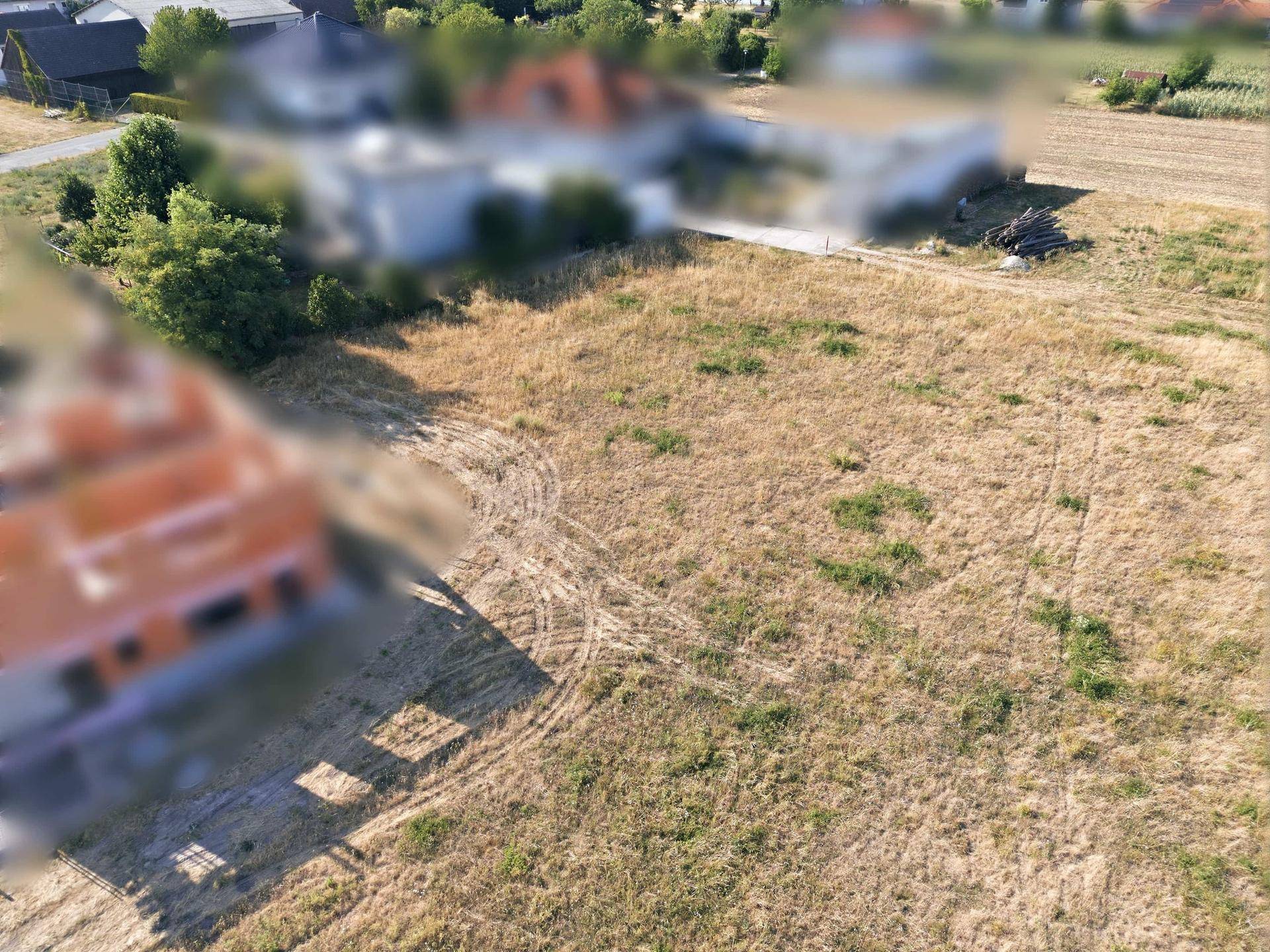 Weitläufiges Feld mit Spuren im Gras und verschwommenen Wohngebäuden in der ländlichen Umgebung.