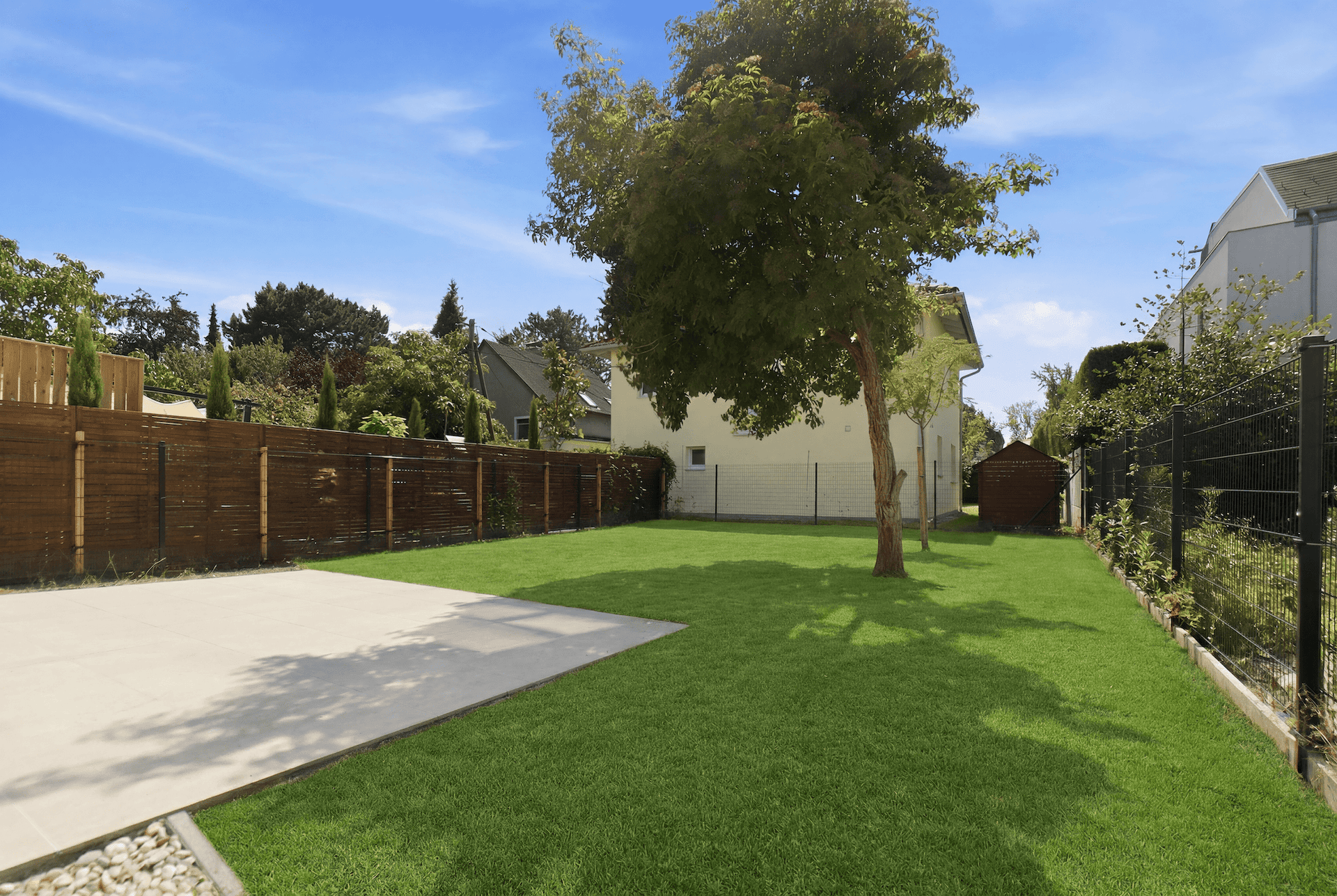 Gepflegter Garten mit Rasenfläche, Baum, gepflastertem Bereich und Holzzaun.
