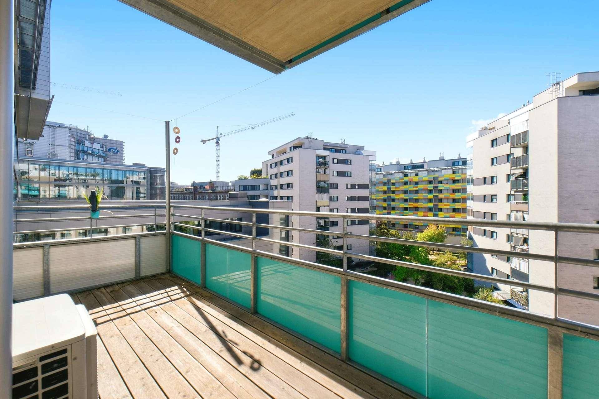 Sonniger Balkon mit Holzdielen und Blick auf die moderne Stadtarchitektur.